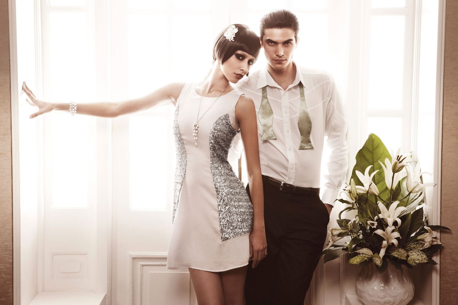 Couple in elegant white outfits pose near window with floral arrangement in bright airy interior.