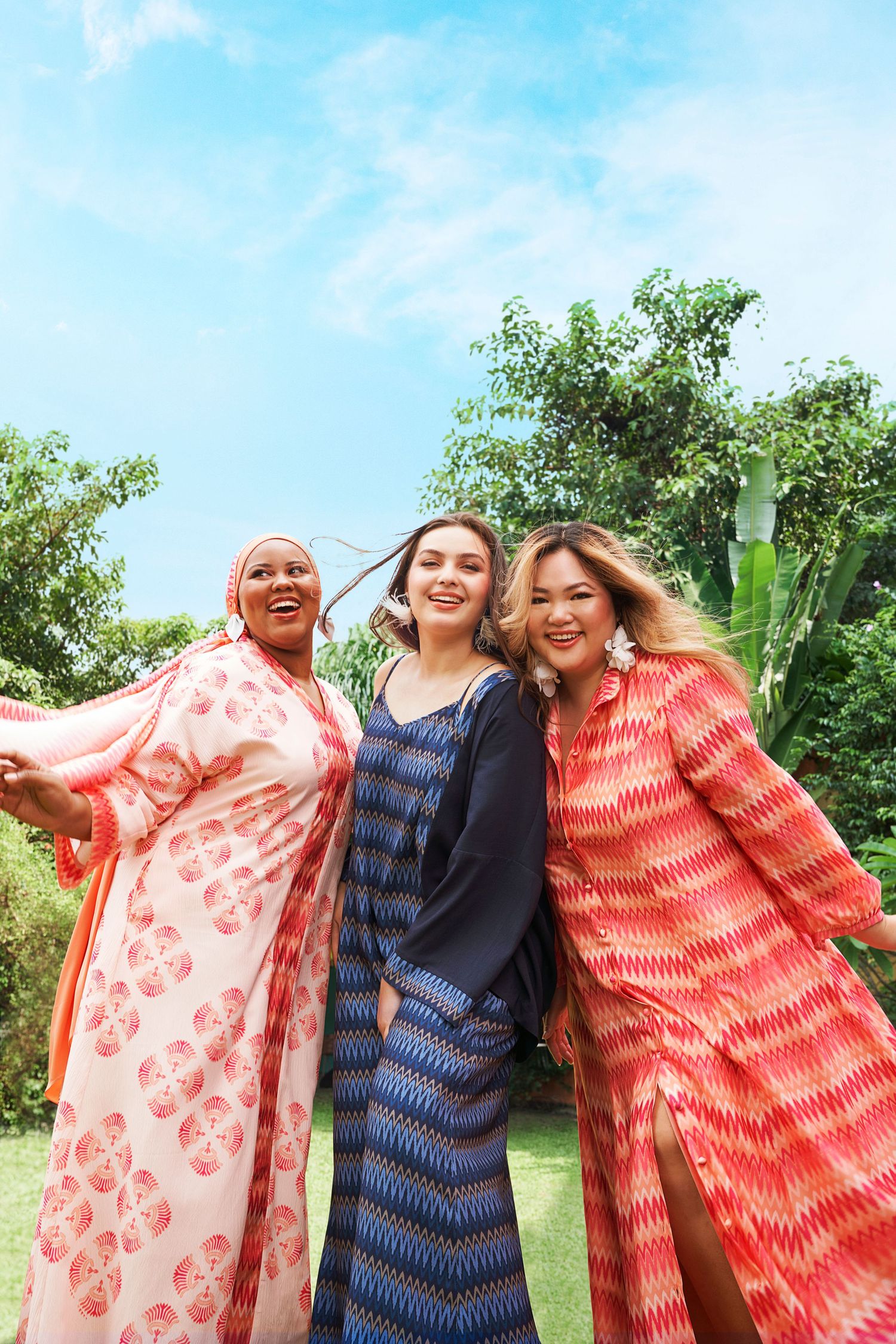 Three diverse plus-size women in vibrant printed maxi dresses posing together in a lush garden.