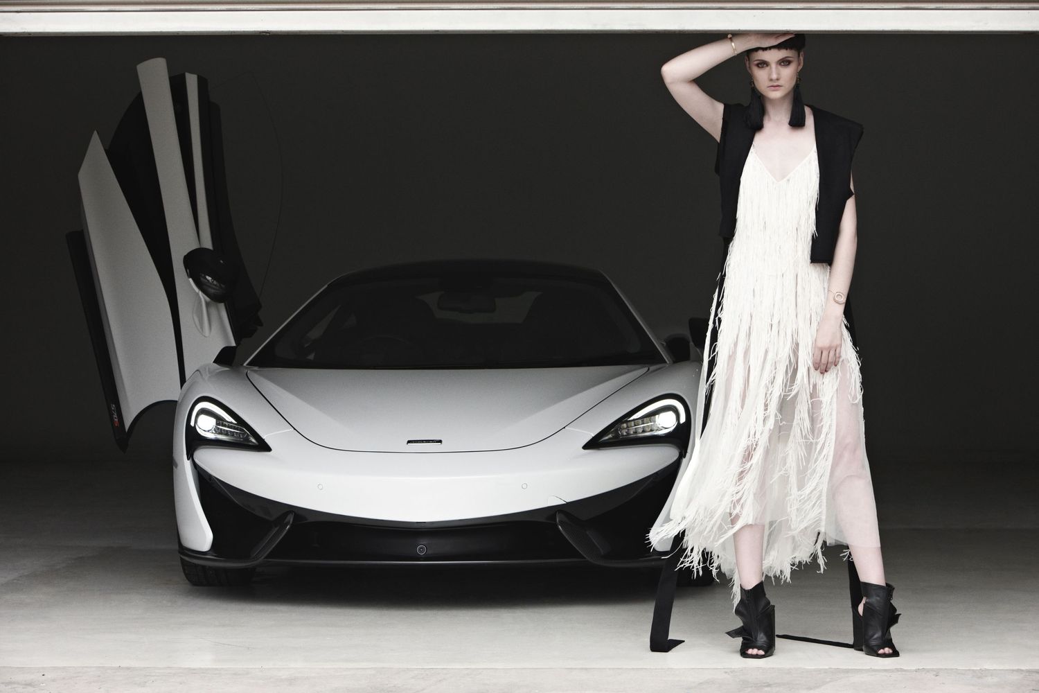 A model in a white fringe dress poses beside a silver McLaren sports car with open door.