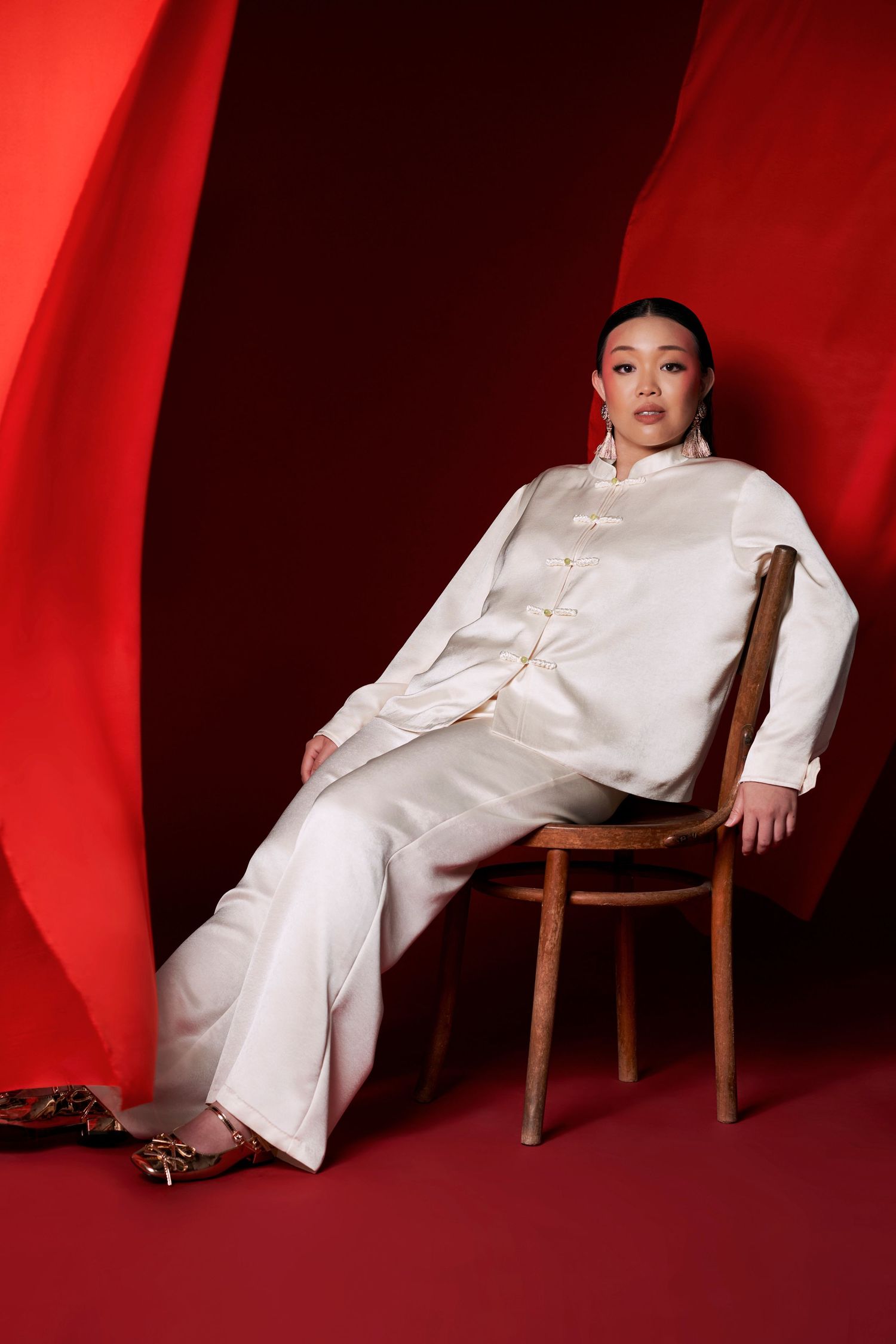 Asian woman in white satin mandarin collar pantsuit sits relaxed on wooden chair with red backdrop.