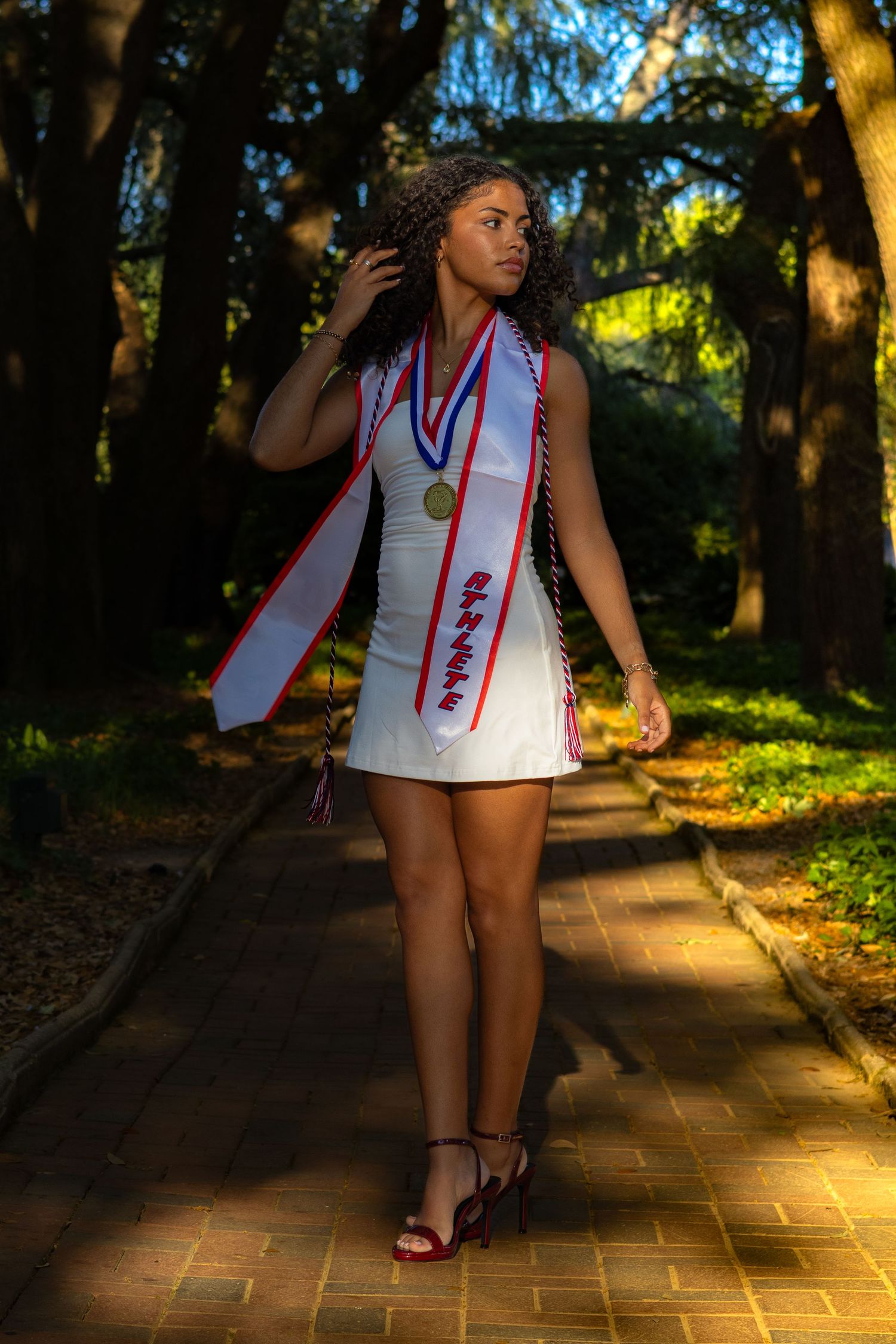 Young woman in white graduation dress with sash and medal walks a sunlit brick path through lush green trees.