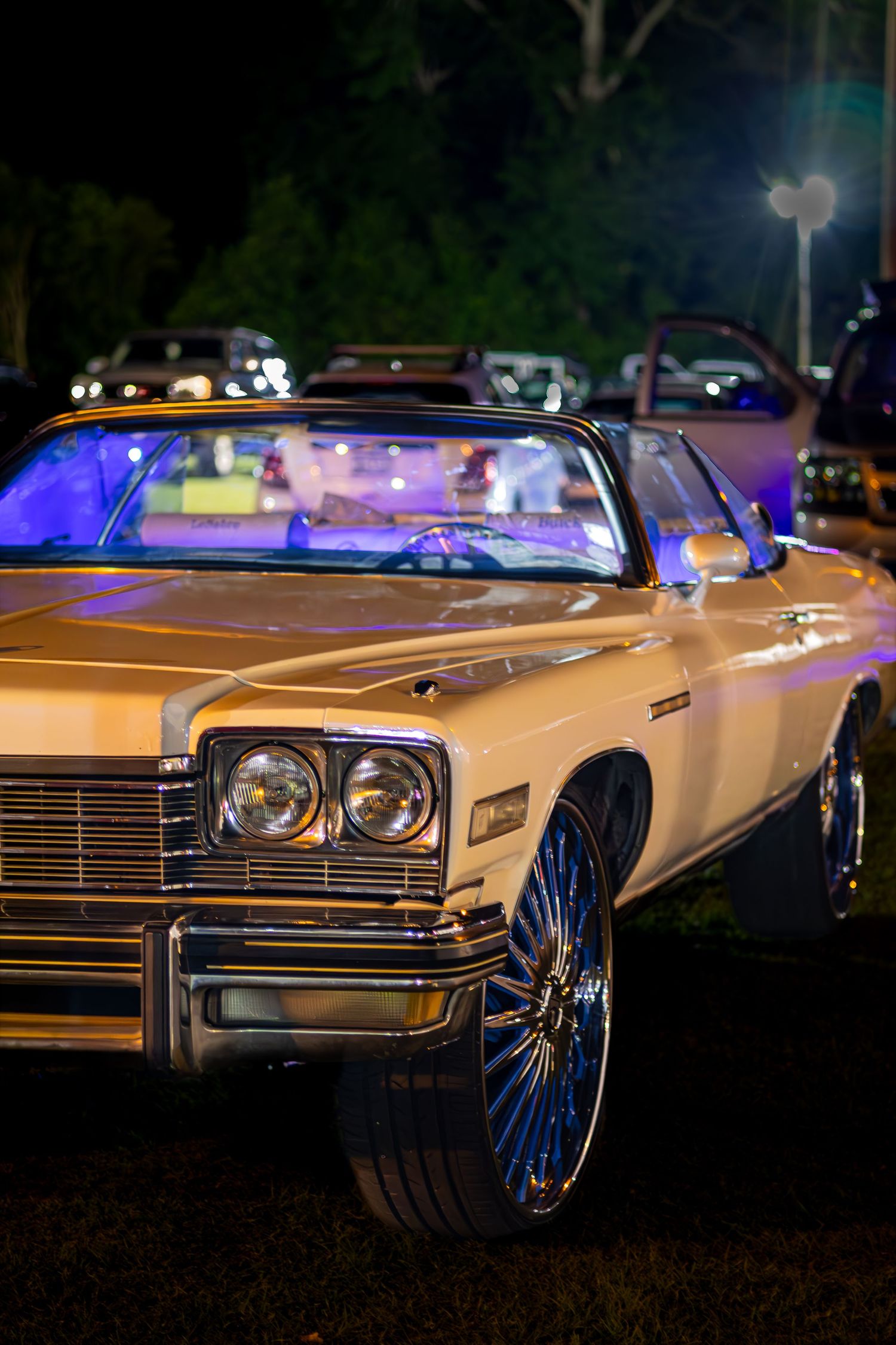 Gold classic convertible lowrider with chrome wheels and purple interior lighting displayed at a nighttime car show.