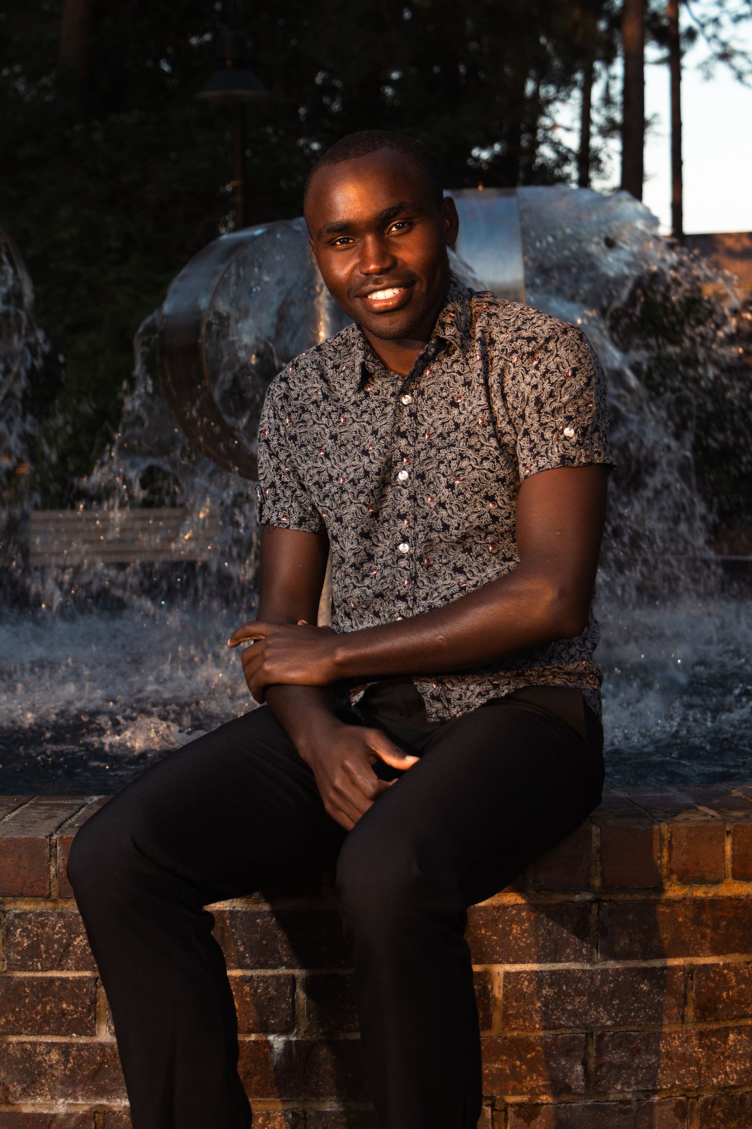 Smiling young man in patterned shirt sitting on brick fountain ledge outdoors at dusk.