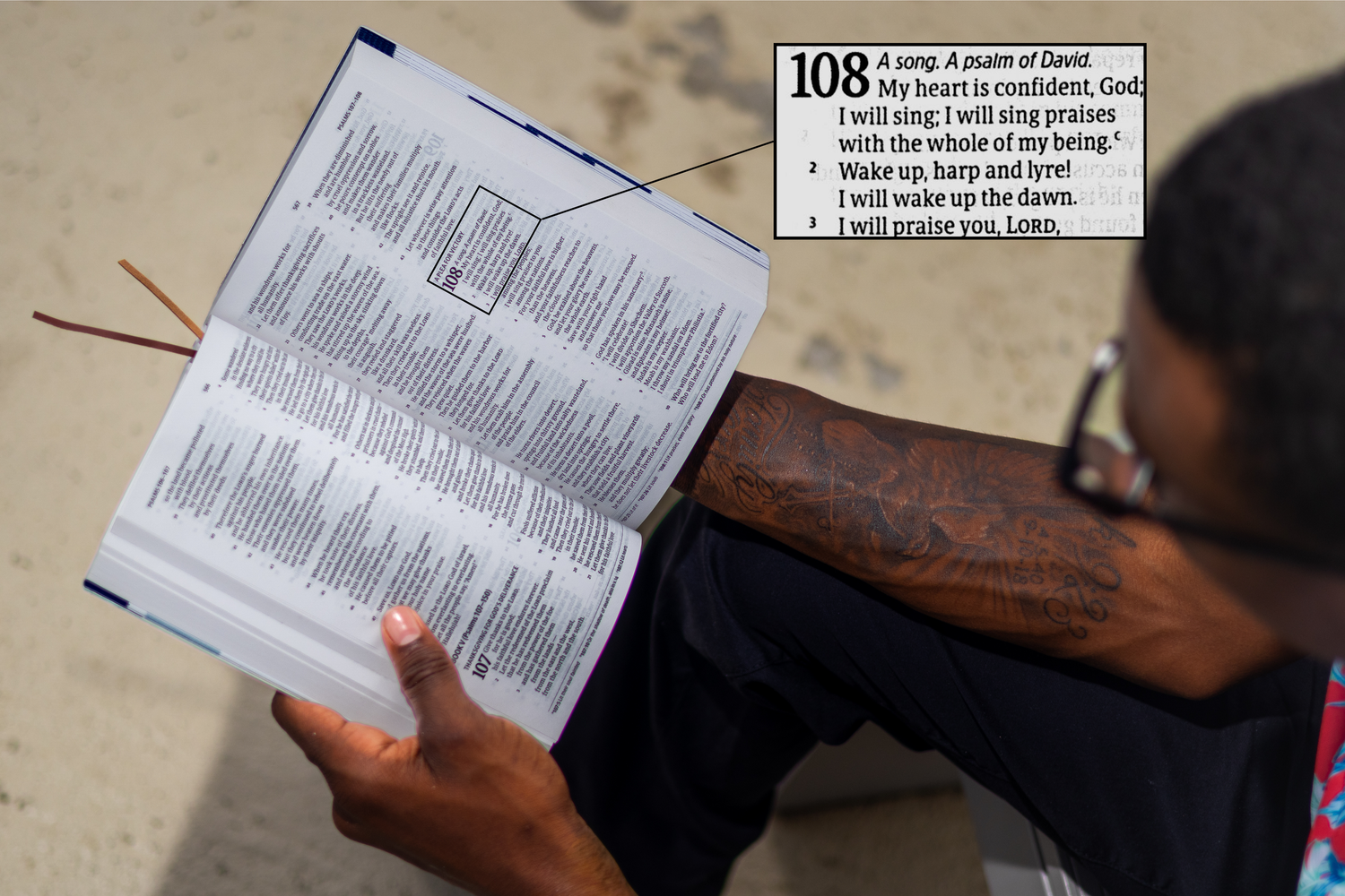 Person with tattoos reading Psalm 108 in an open Bible outdoors in natural light.