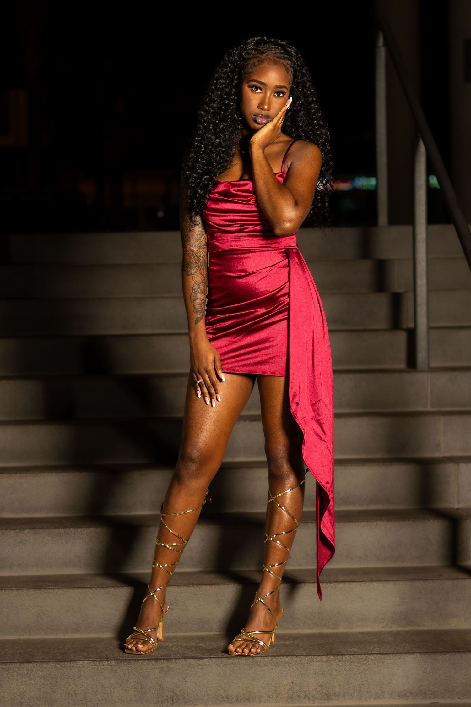 Woman in red satin mini dress with gold strappy heels posing on stairs at night.