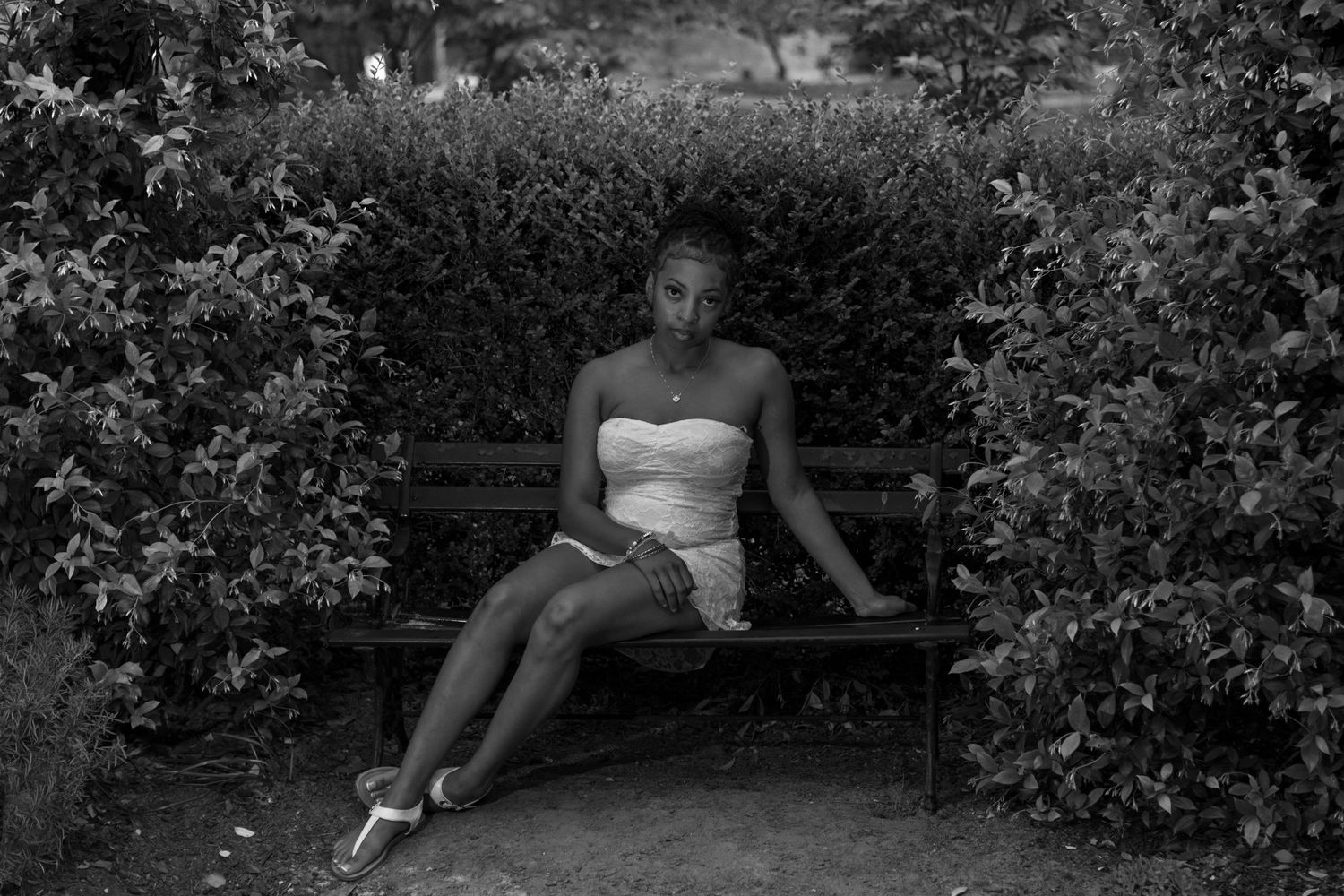 Black and white photo of a young woman in a strapless dress sitting on a park bench surrounded by lush hedges.
