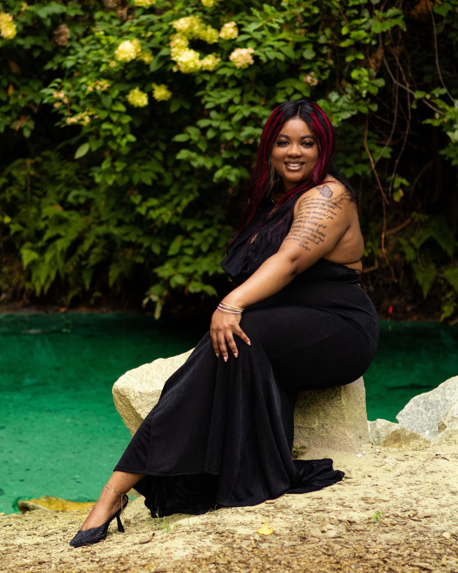 A woman in a black outfit sits on a rock near a green pond, surrounded by lush yellow flowering bushes.