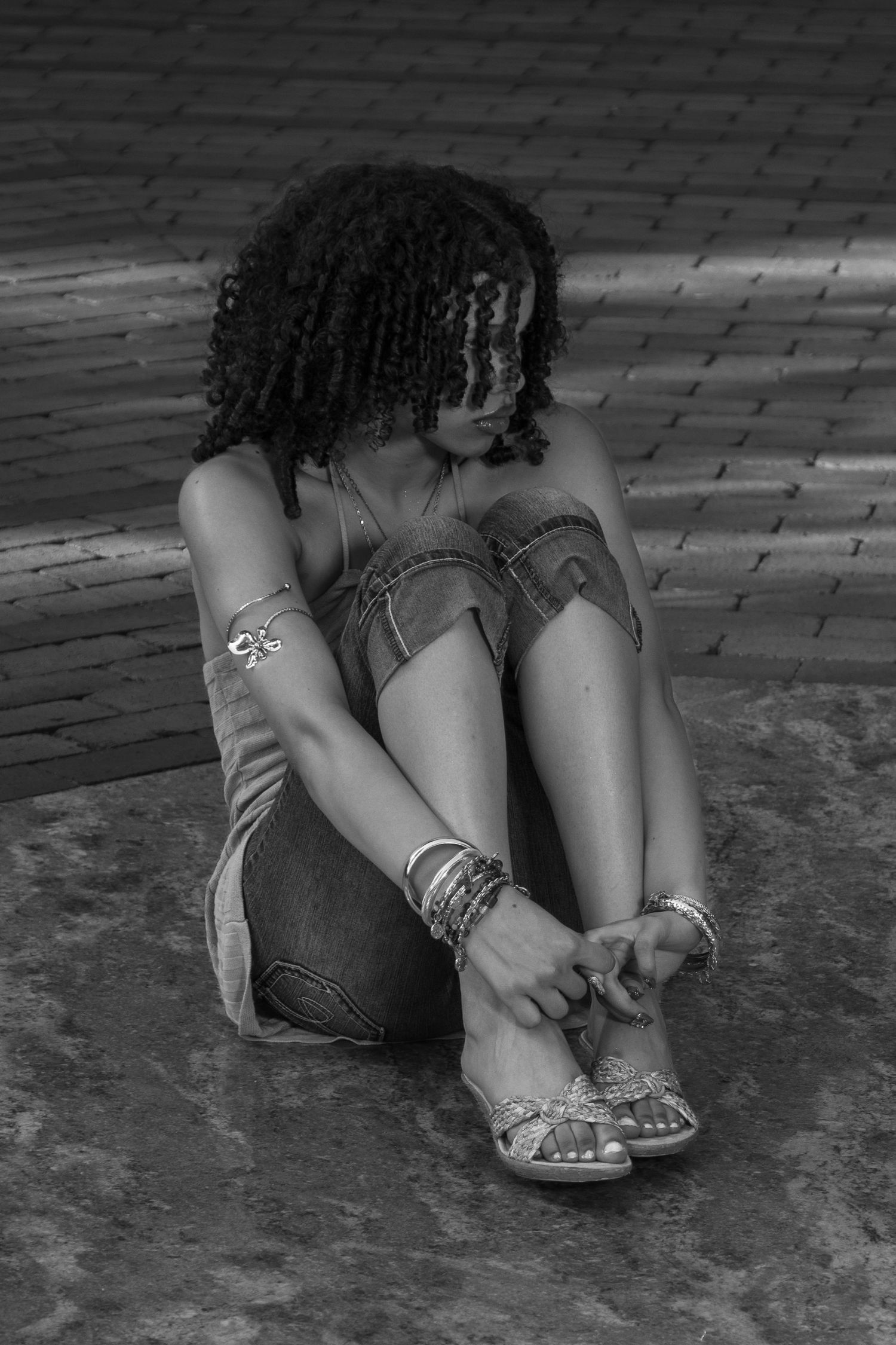 Black and white photo of young woman adjusting sandal while sitting on brick ground.