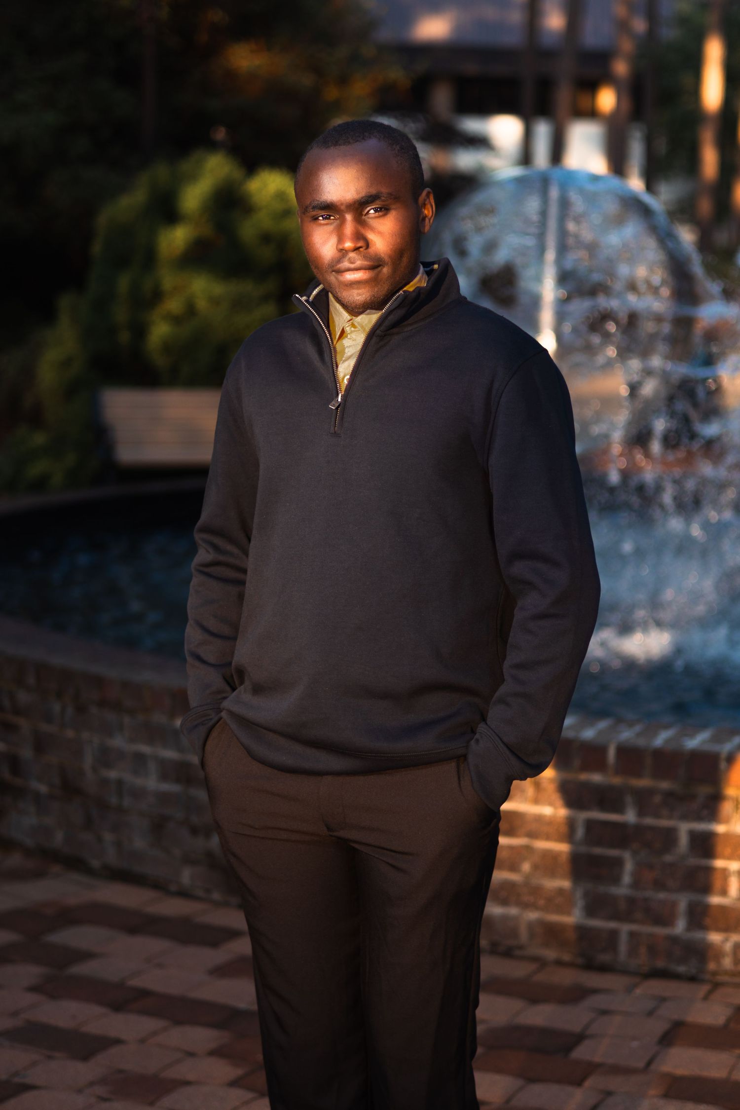 Young man in black quarter-zip sweater stands confidently near a decorative fountain at dusk.