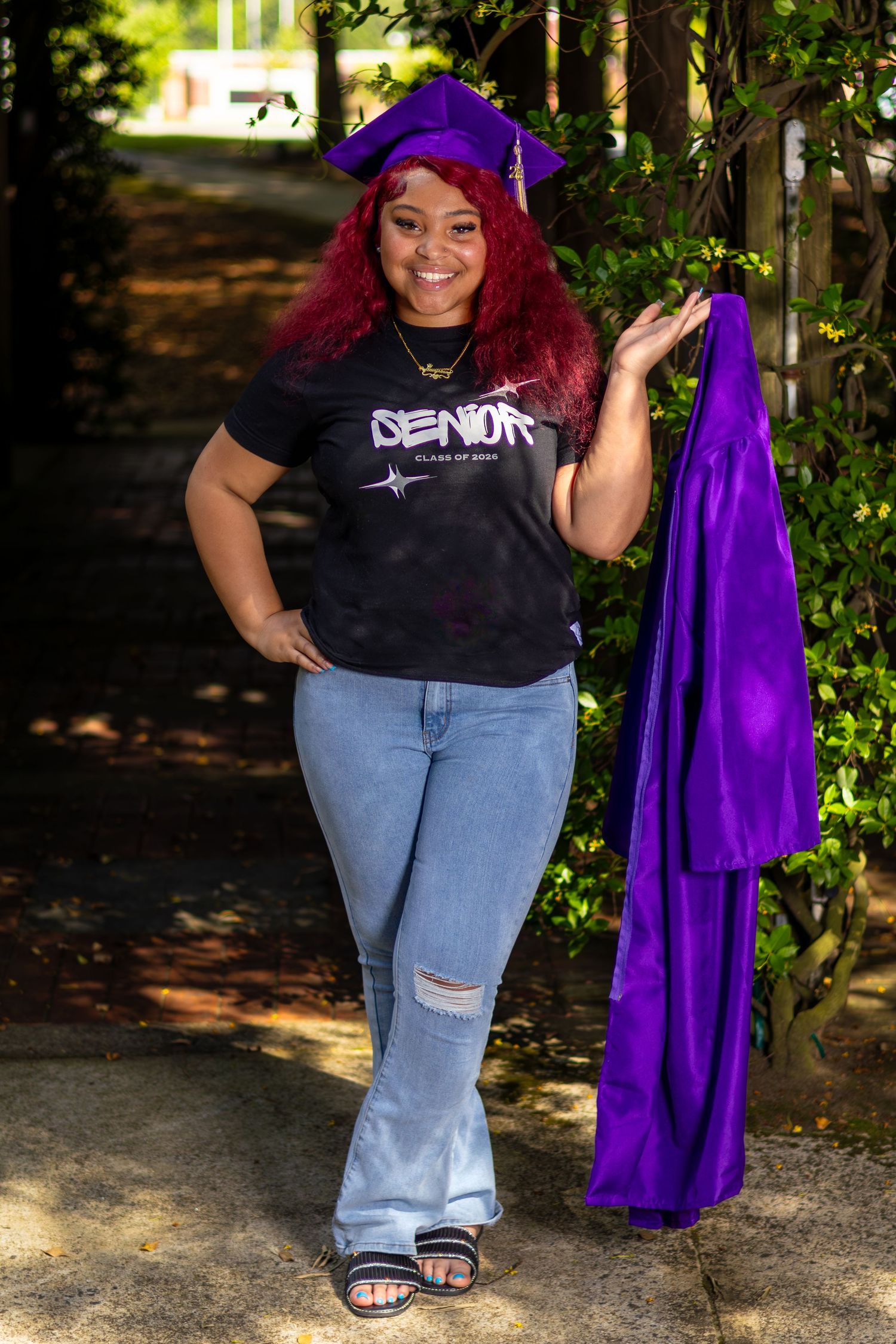 Young woman in purple graduation cap holding gown, wearing Senior shirt and jeans, smiling outdoors.