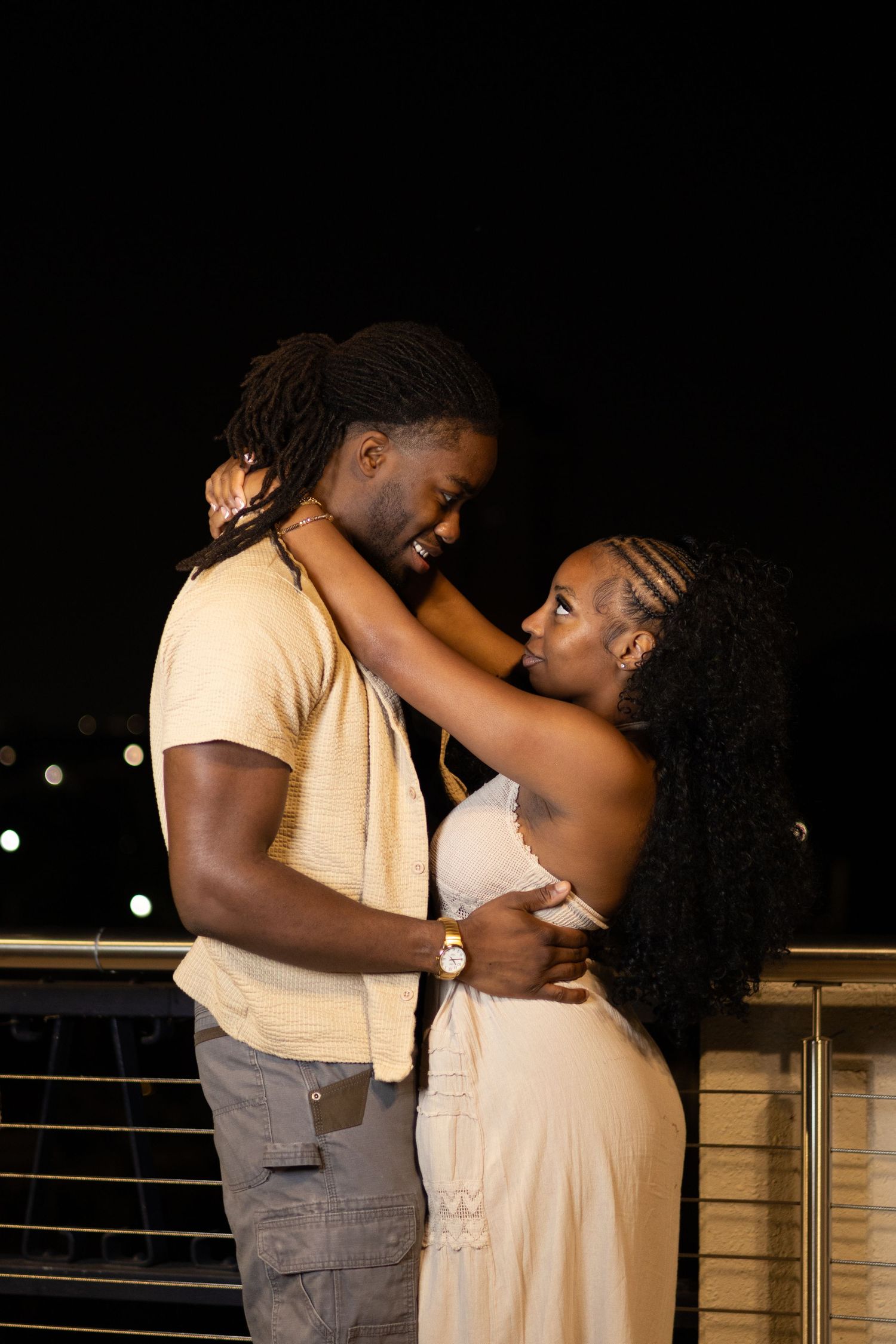 A romantic Black couple embraces and gazes into each other's eyes on a balcony at night.