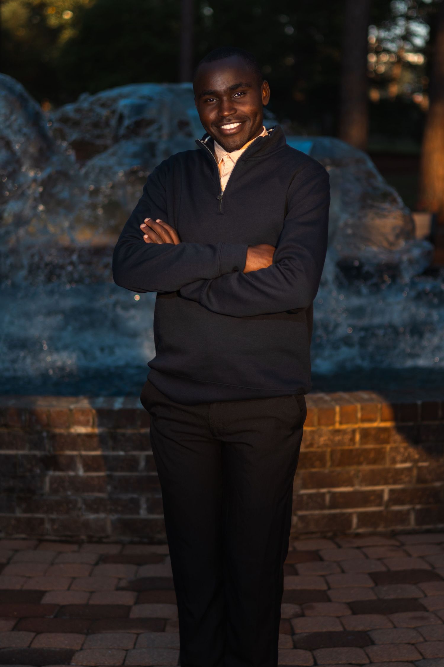 Smiling man in black sweater stands arms crossed before illuminated fountain at dusk.