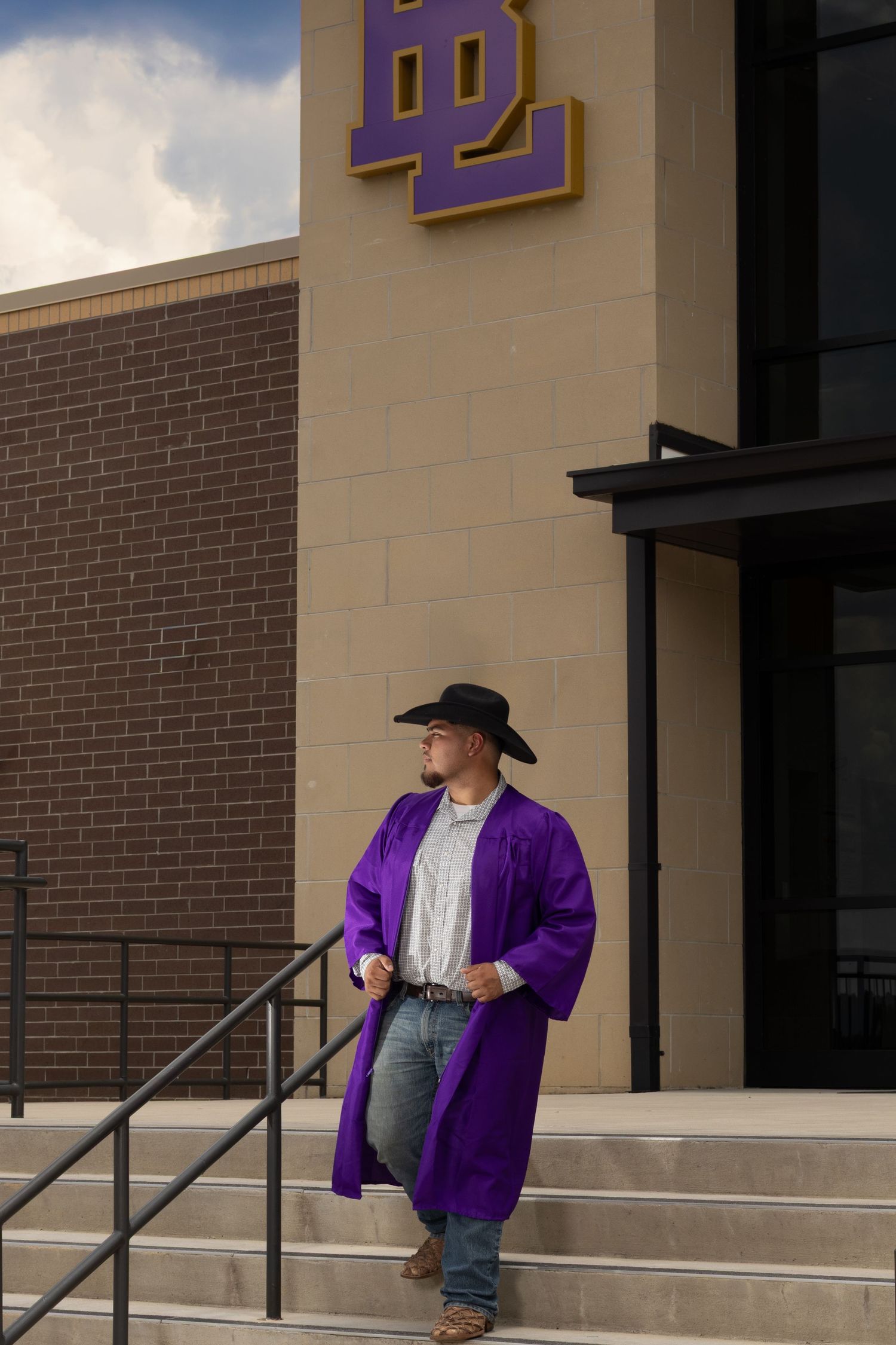 A young man in a purple graduation gown and black cowboy hat stands on steps outside a school building with purple and gold logo.