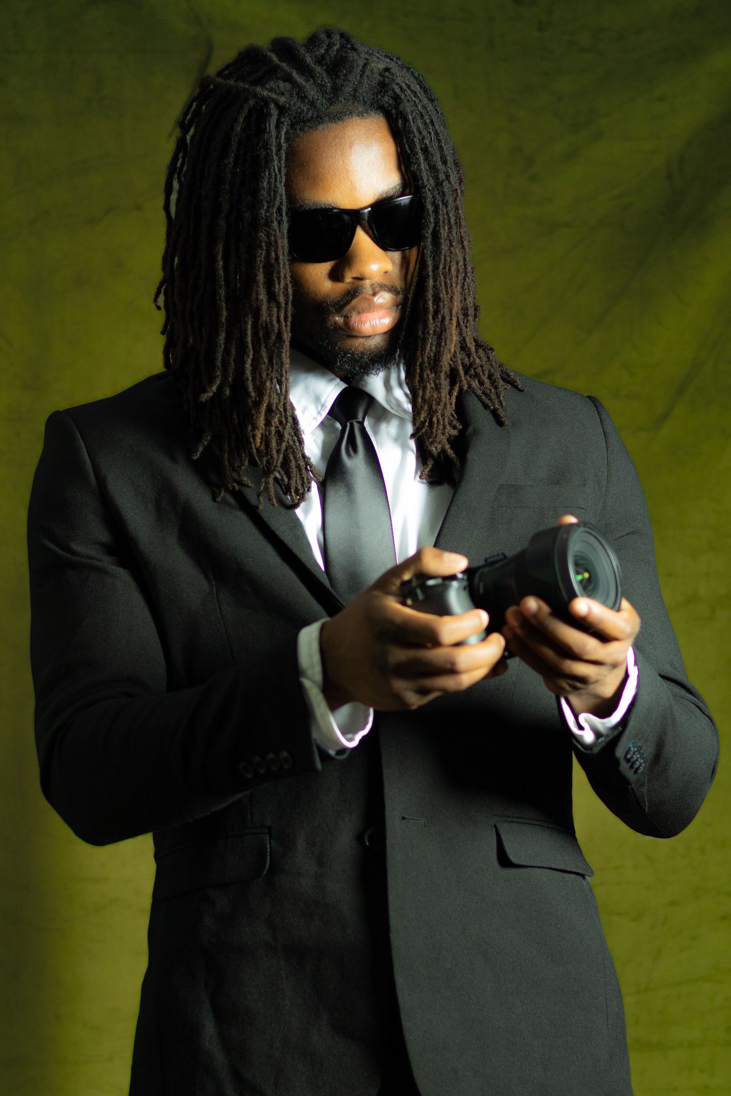 Man in black suit with dreadlocks and sunglasses holding a camera against a green background.