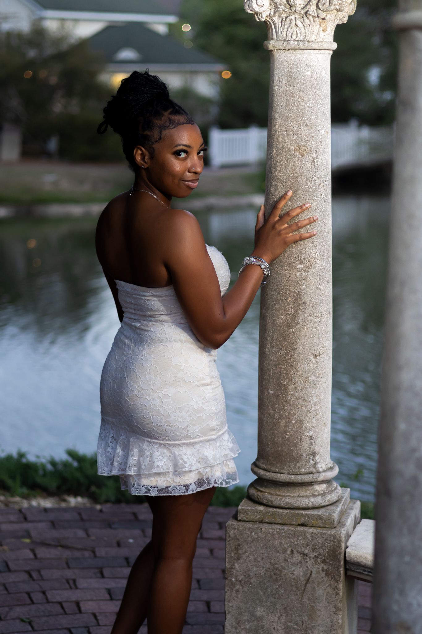 Young woman in white lace dress posing elegantly by a stone column near a serene waterfront at dusk.
