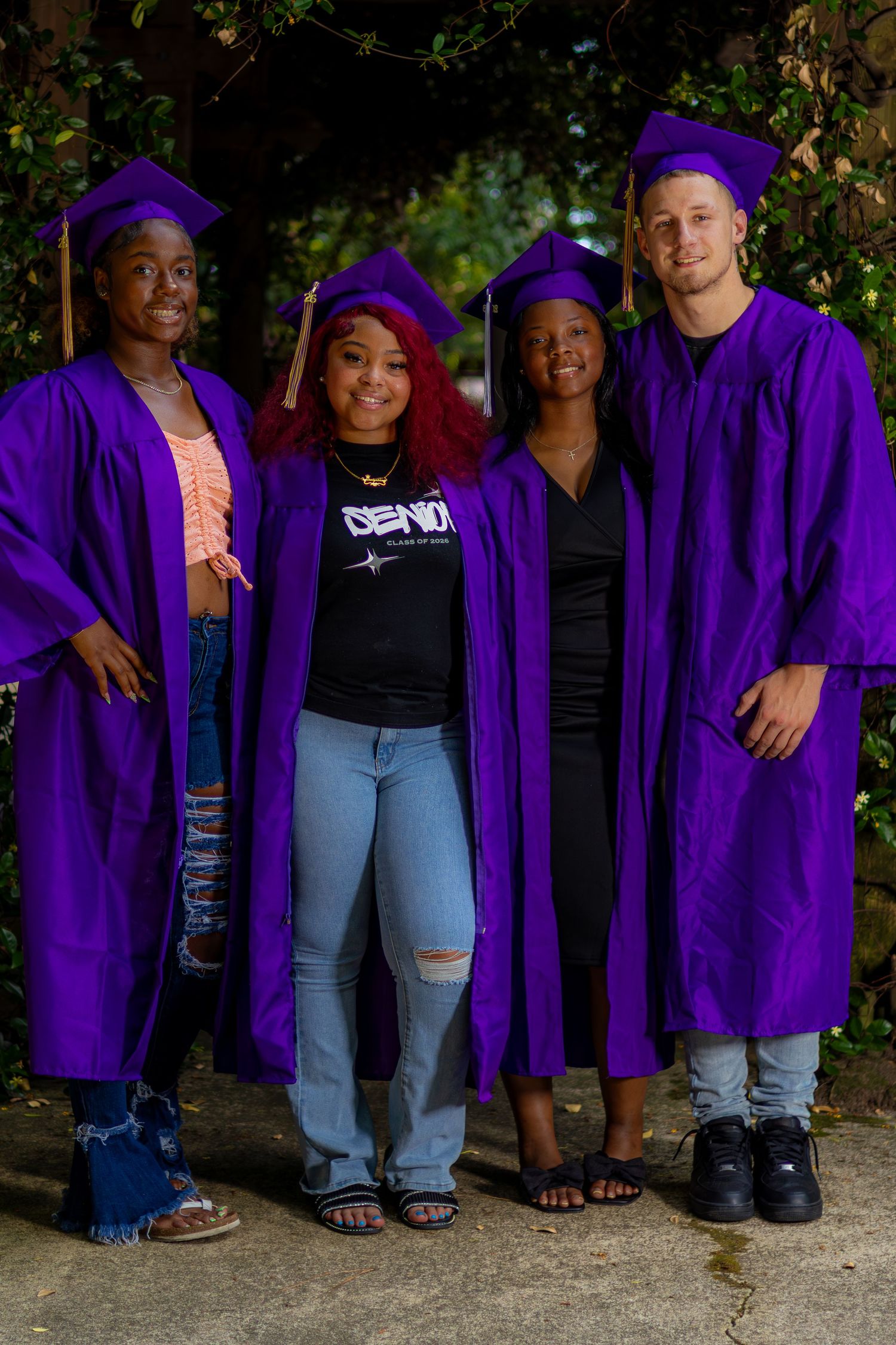 Four high school seniors in purple graduation caps and gowns posing together outdoors for a graduation photo.