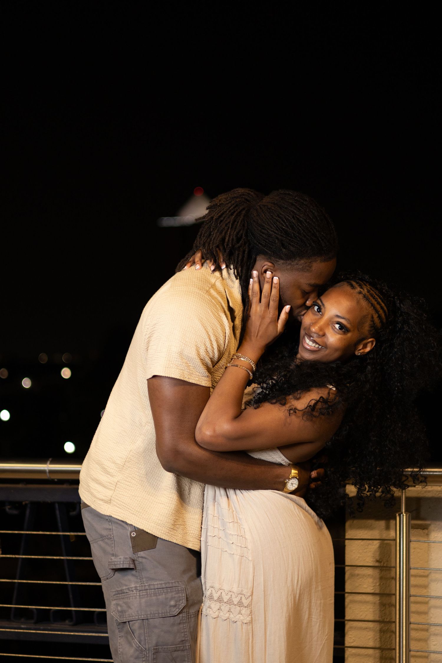 A couple shares a romantic moment at night, the man kissing the smiling woman's cheek on a city balcony.