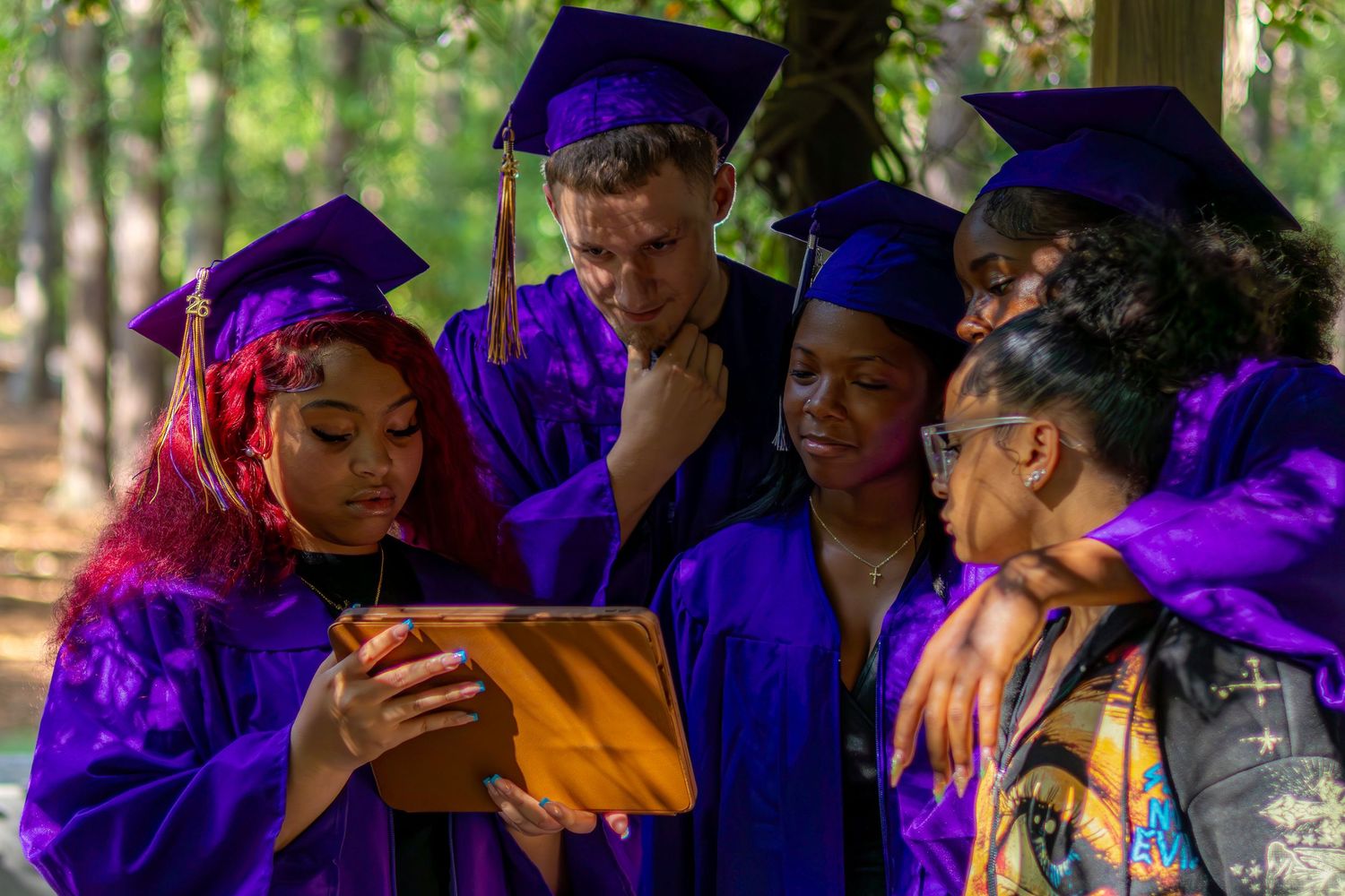 Group of graduates in purple caps and gowns viewing a tablet together outdoors among trees.