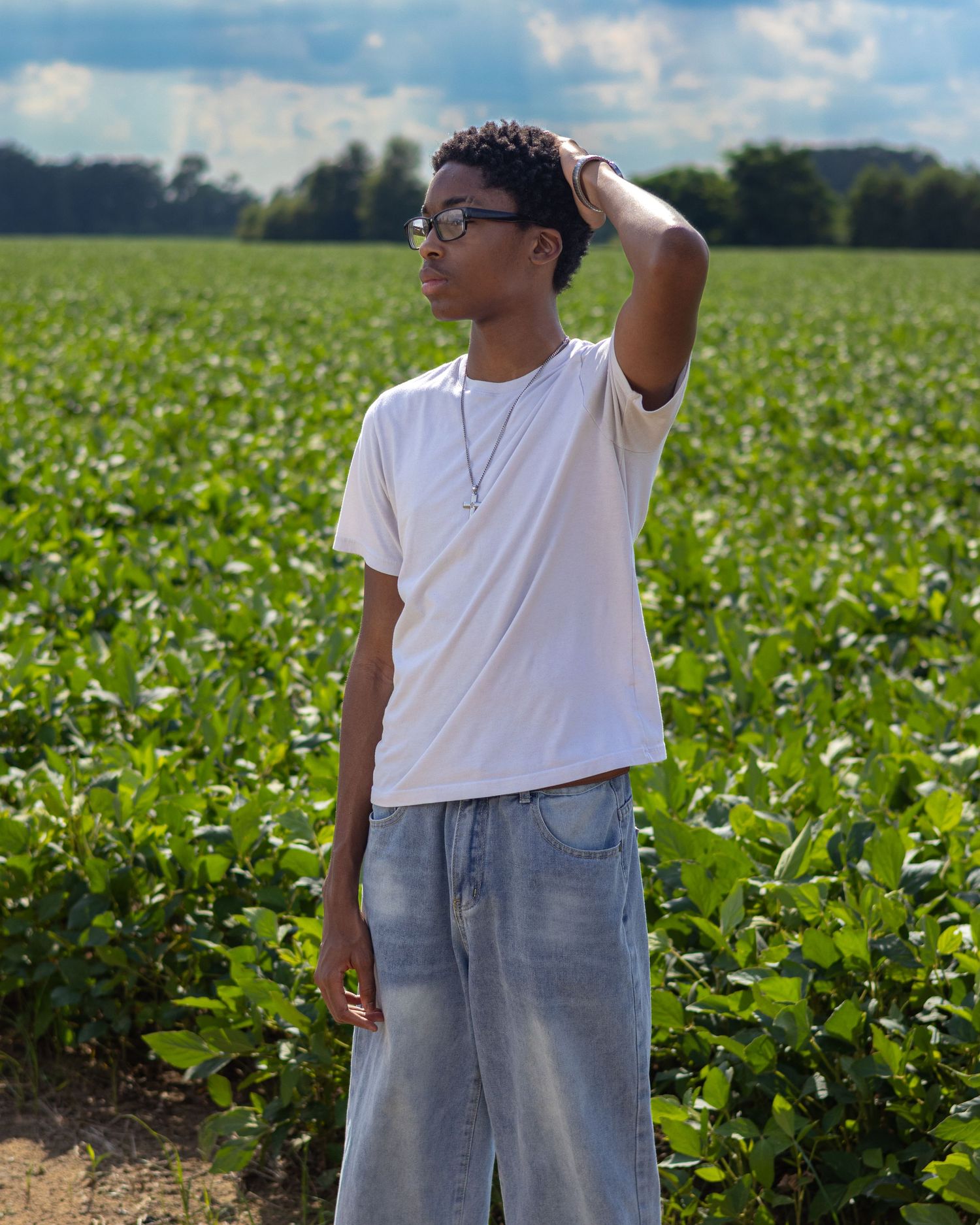 Young man in white tee and baggy jeans poses in lush green farm field, hand on head, wearing glasses.