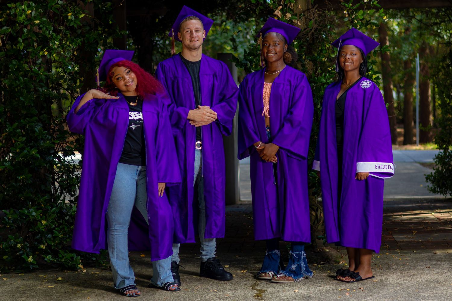 Four graduates in purple caps and gowns pose outdoors on a tree-lined path celebrating their academic achievement.