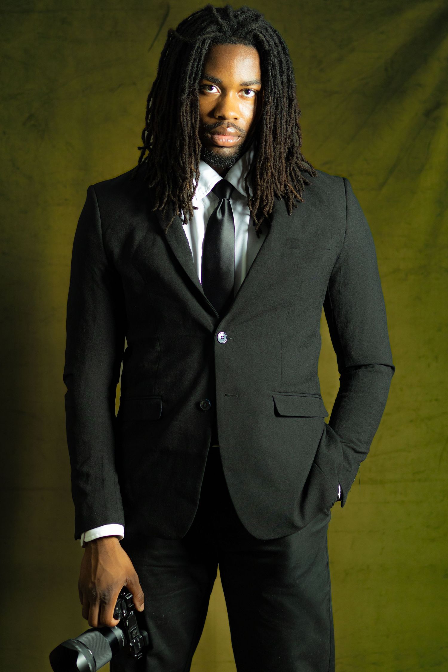Young Black man with dreadlocks in black suit holding camera, posing against olive green background.