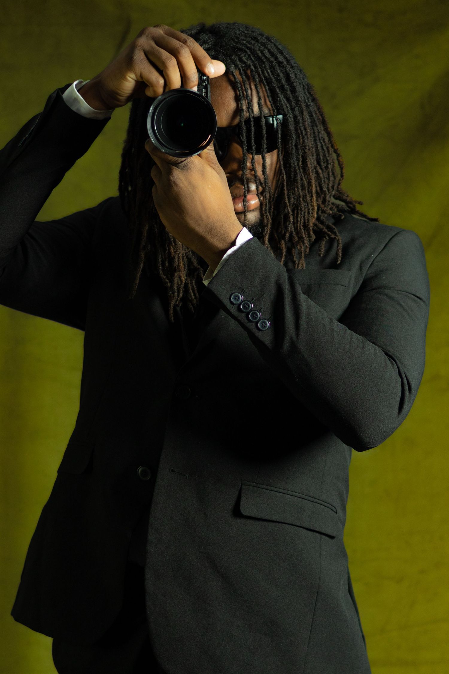 A man in a black suit with dreadlocks holds a DSLR camera up to his face against a yellow backdrop.