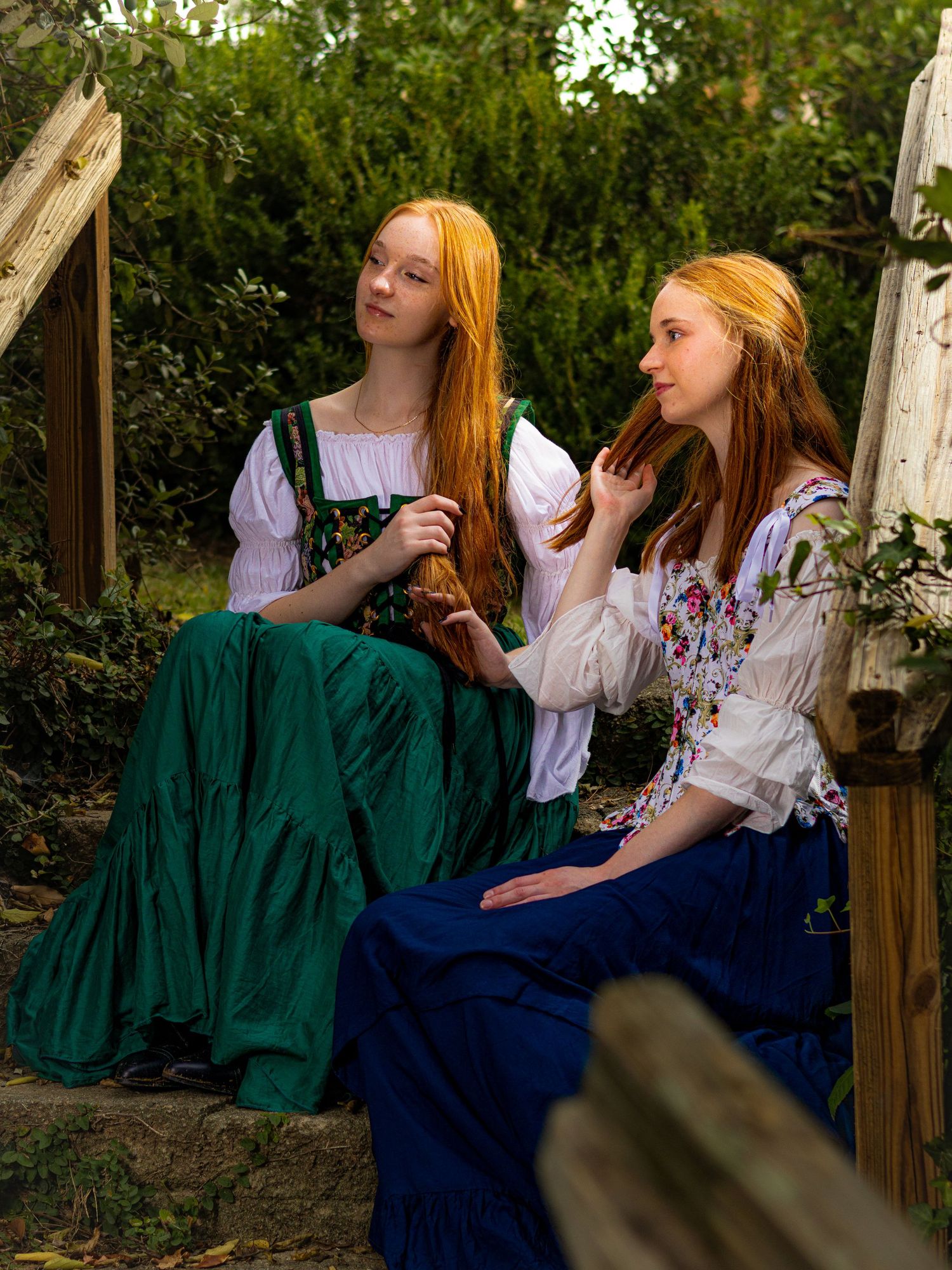 Two red-haired women in Renaissance-style green and blue dresses sit outdoors among lush greenery.