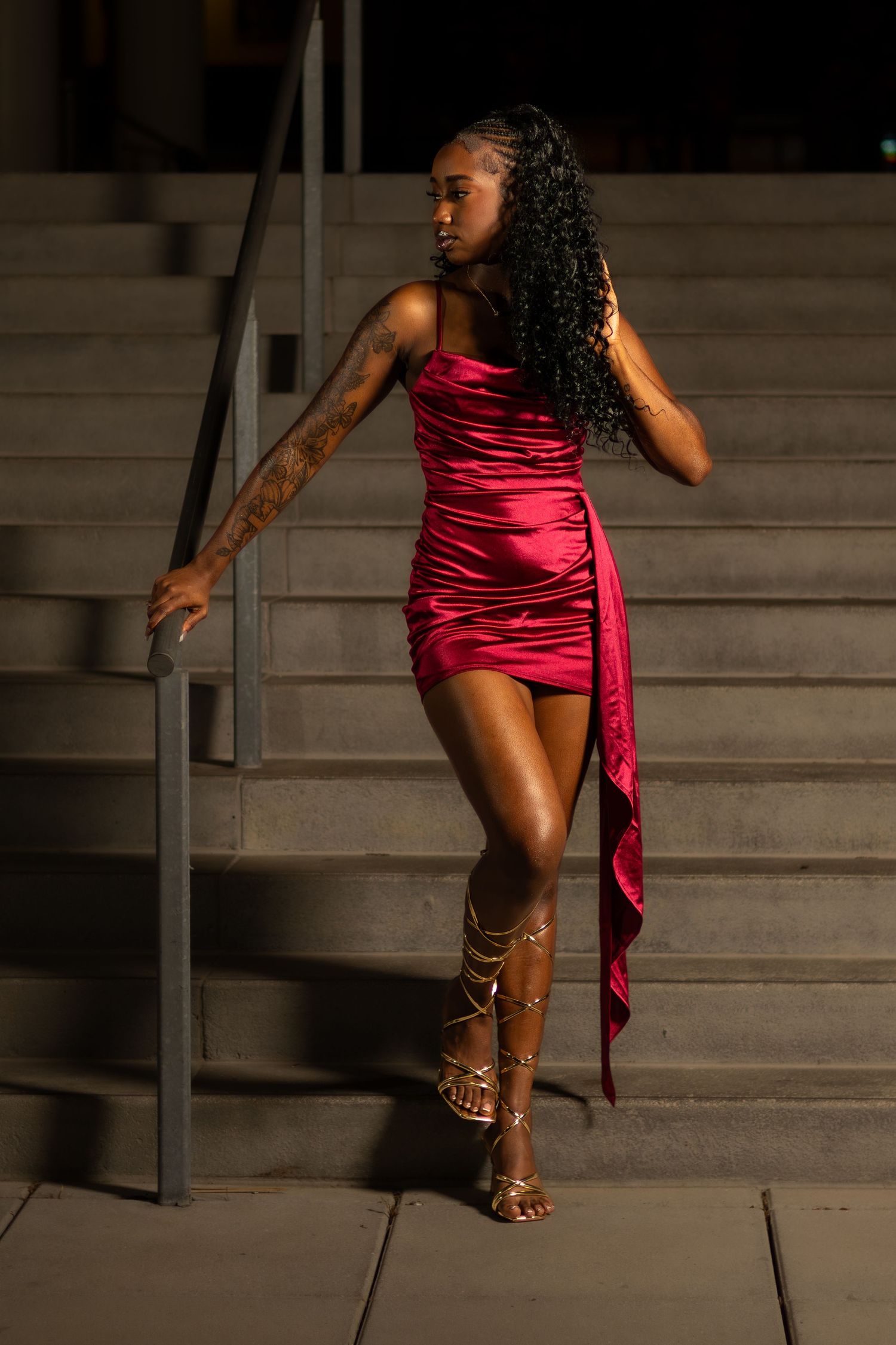 Woman in red satin mini dress and gold gladiator heels poses on concrete steps at night.