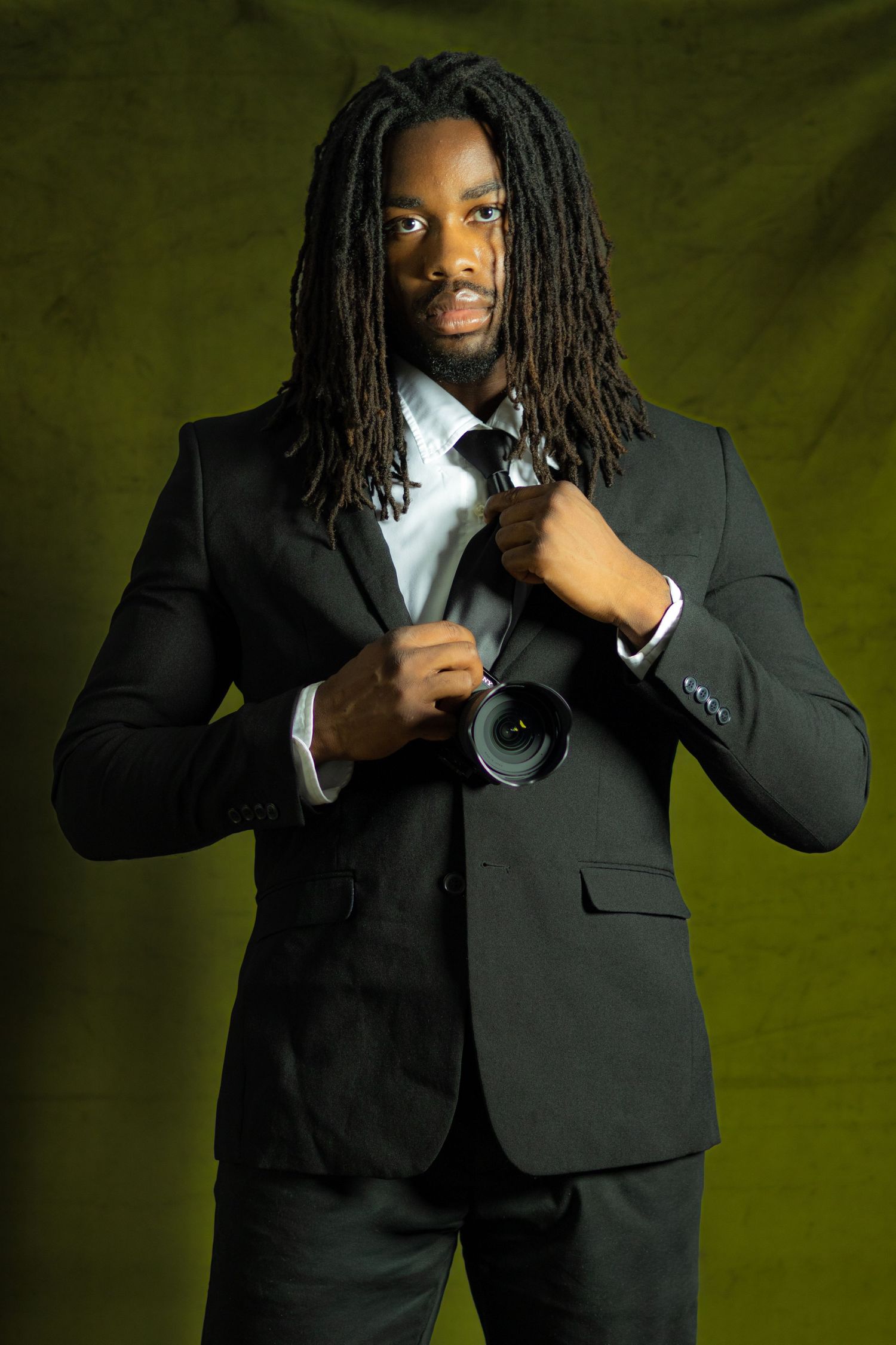 A stylish man with dreadlocks wearing a black suit holds a camera lens while posing against a dark green background.