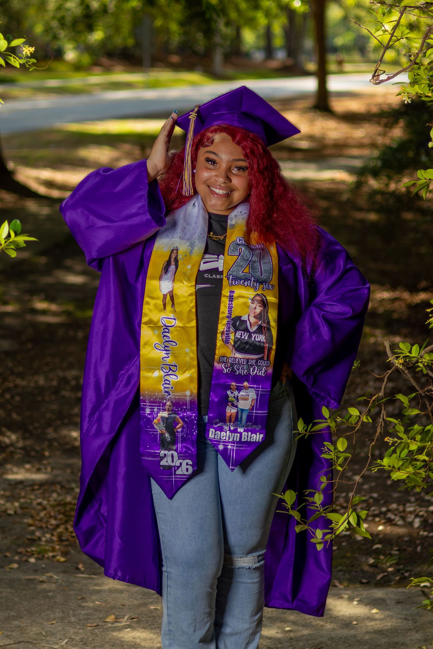 Young woman in purple graduation cap and gown with gold stole smiles outdoors among green trees.