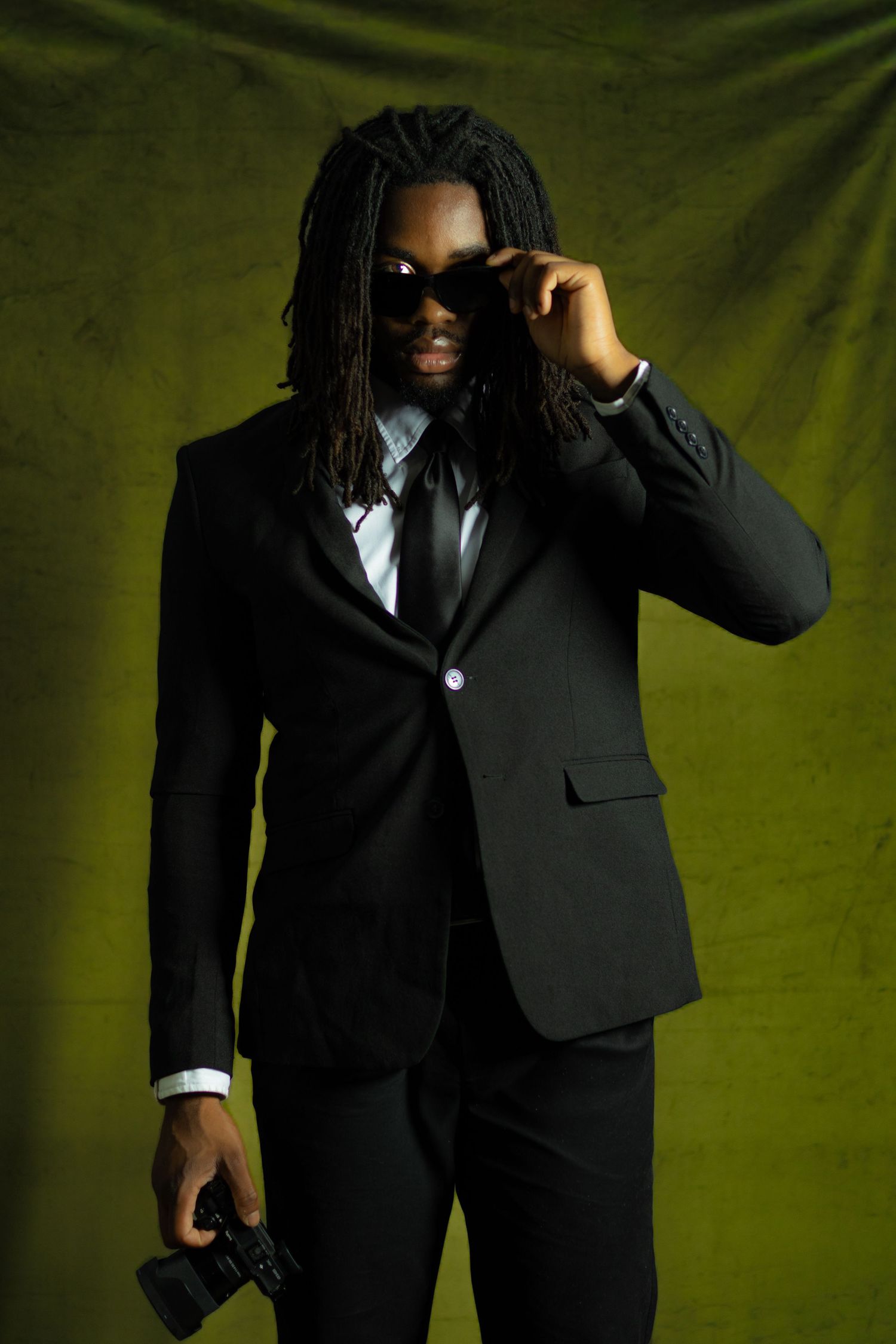 Stylish man in black suit with dreadlocks adjusting sunglasses, holding camera against green backdrop.