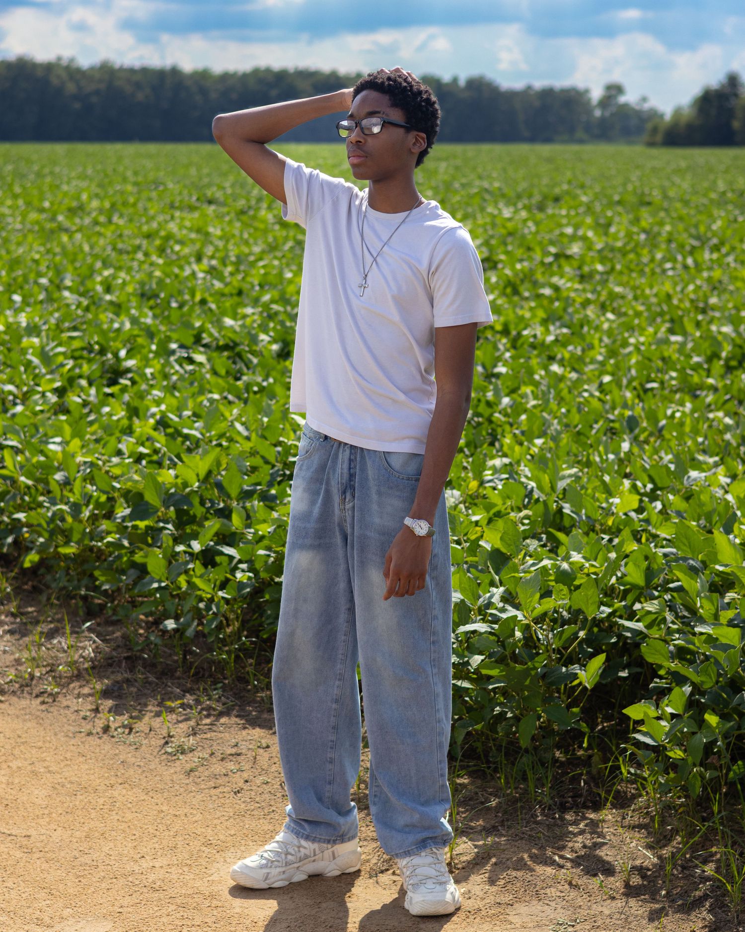 Young man in white tee and light jeans poses on a dirt path beside a lush green crop field on a sunny day.