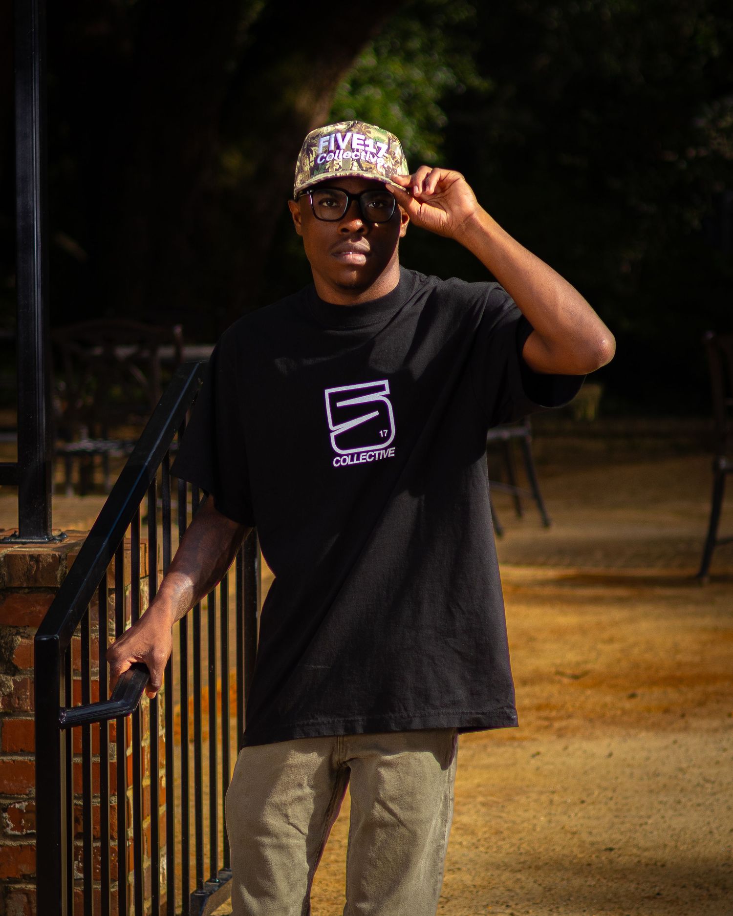 Man in black Five Collective t-shirt and branded cap posing outdoors near iron railing.