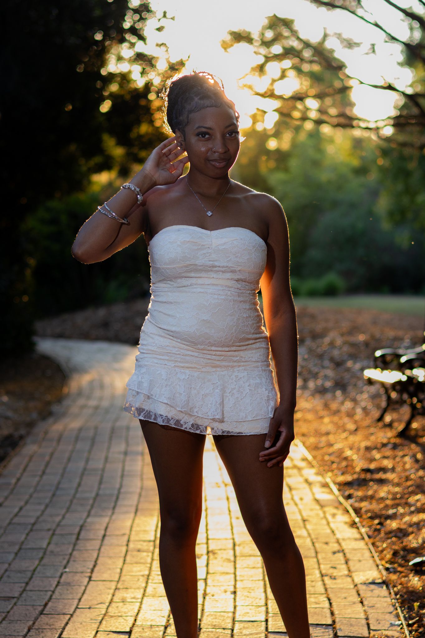 Young Black woman in white strapless mini dress poses on brick path at golden hour in a lush green park.