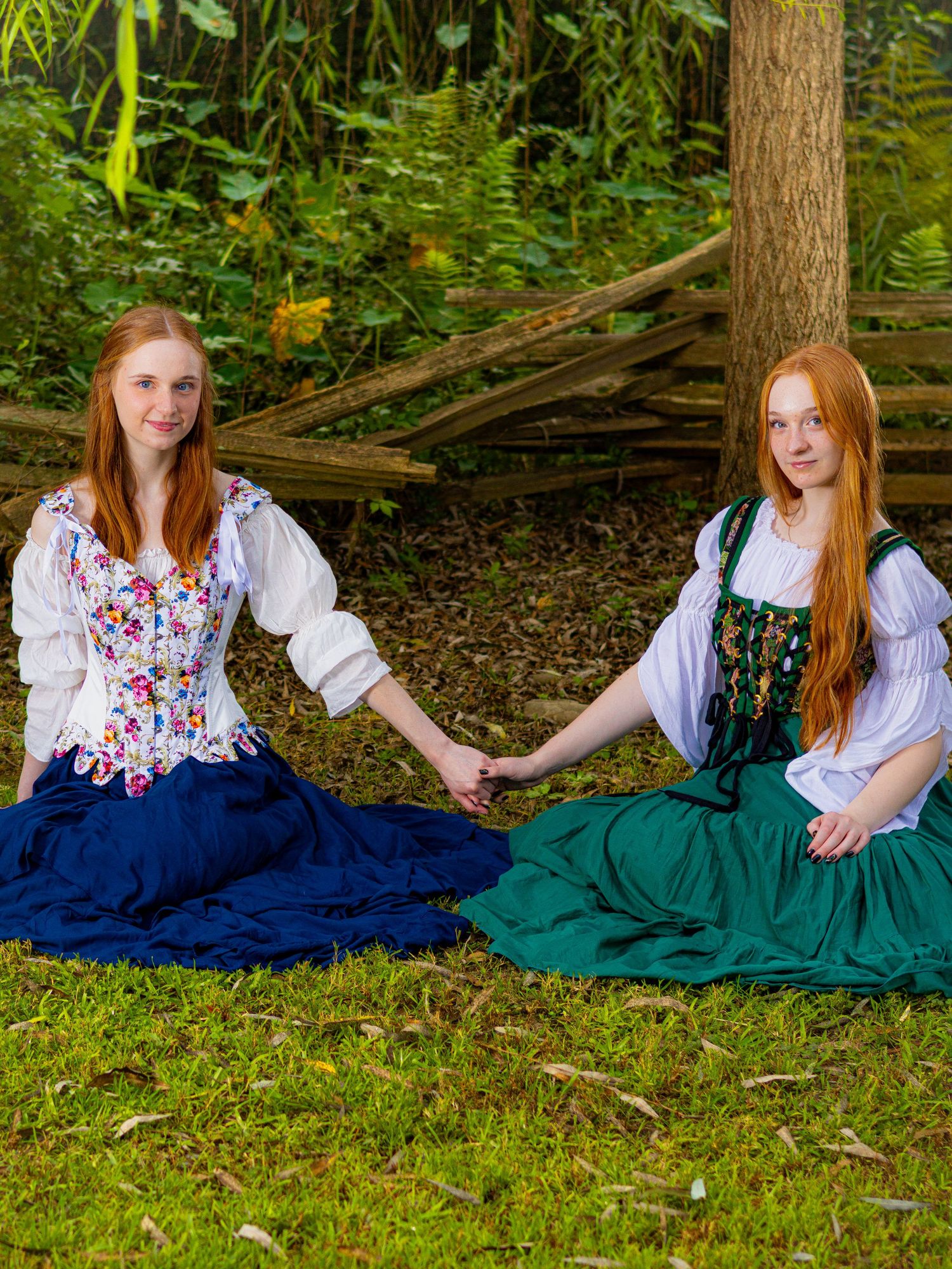 Two redhead women in medieval Renaissance dresses sit on grass holding hands near a wooden fence outdoors.