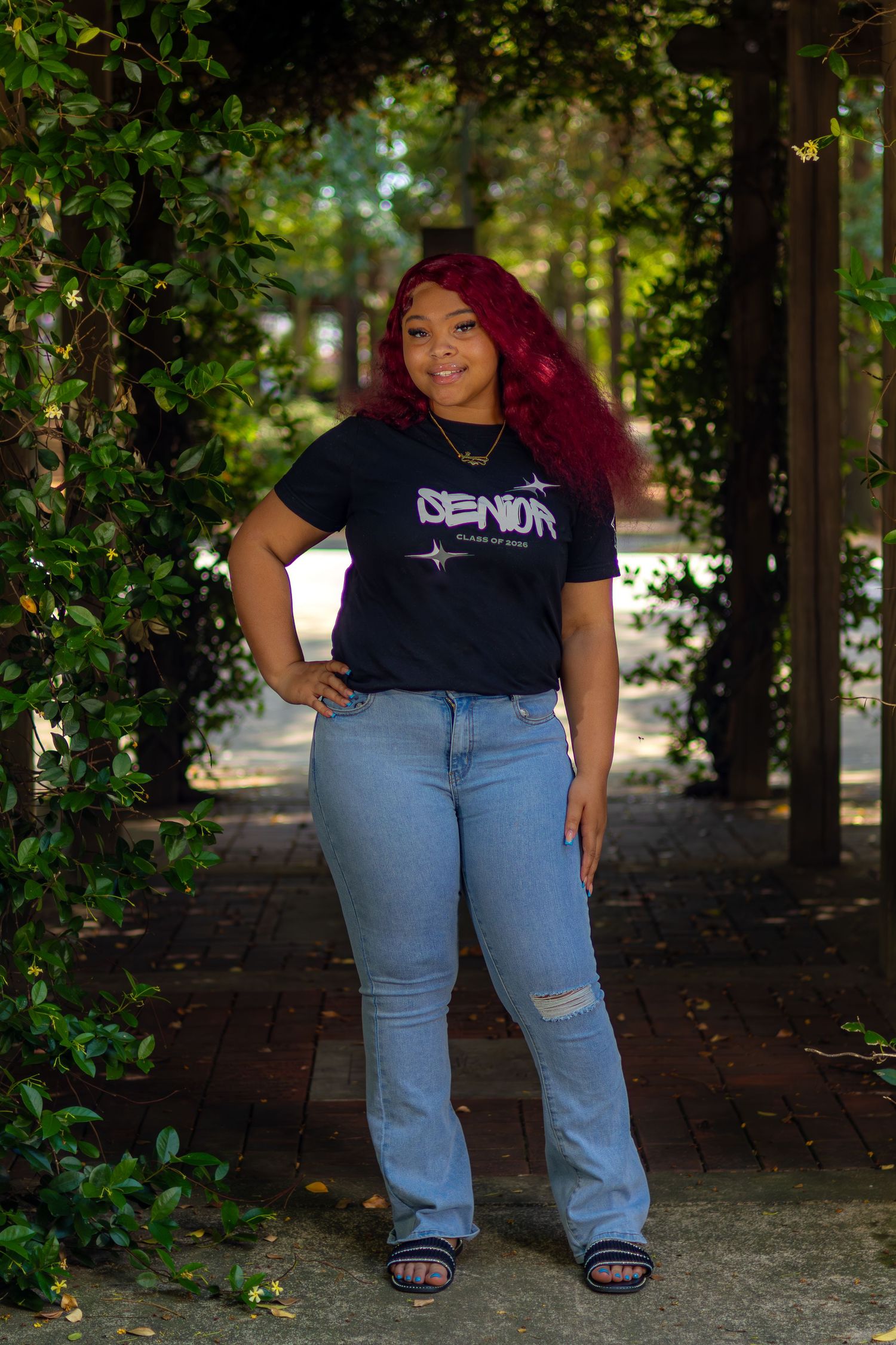 Young woman in black Senior shirt and light blue jeans poses under vine-covered archway on brick path.