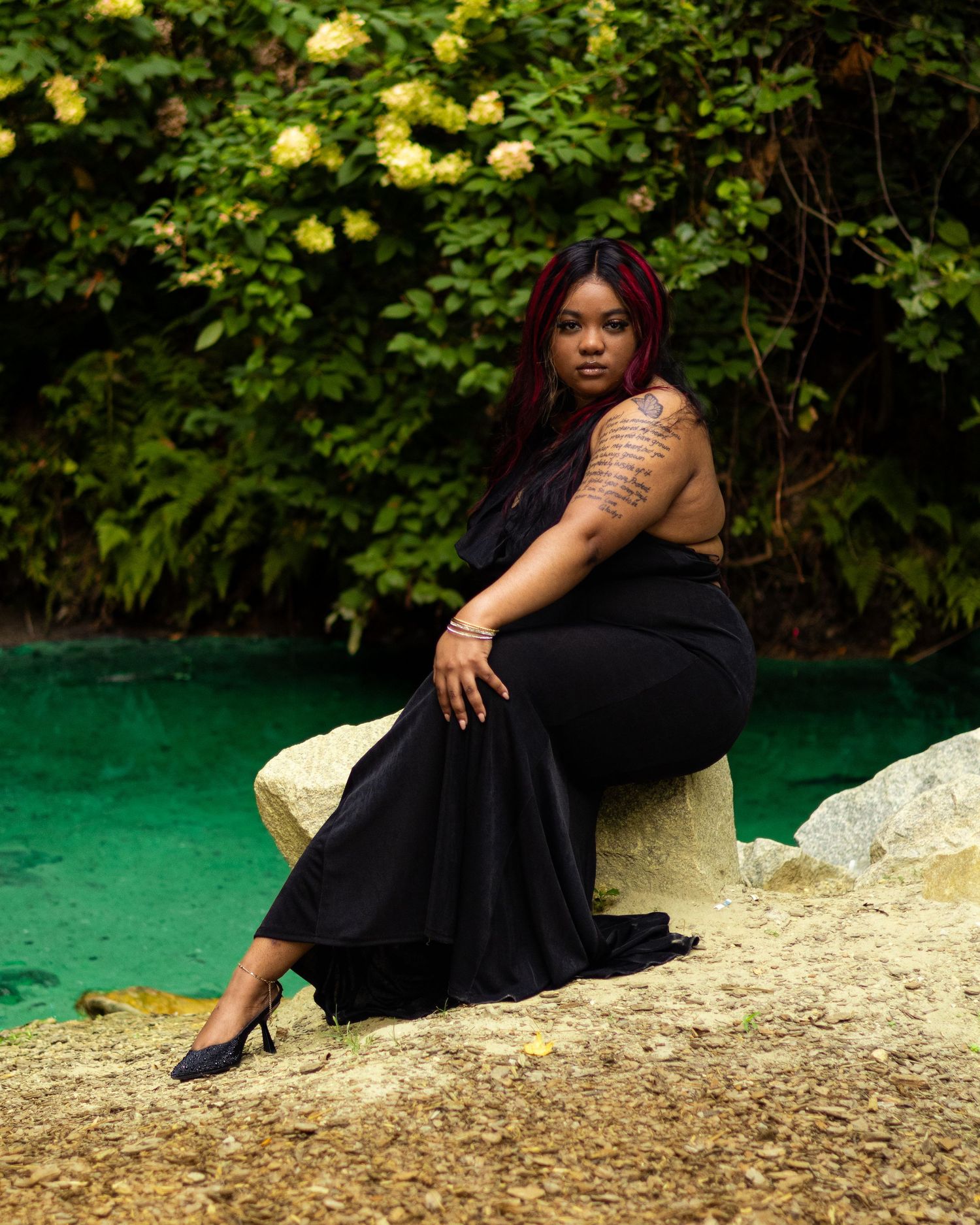 Woman in black dress sitting on rocks by turquoise water, surrounded by lush green foliage.