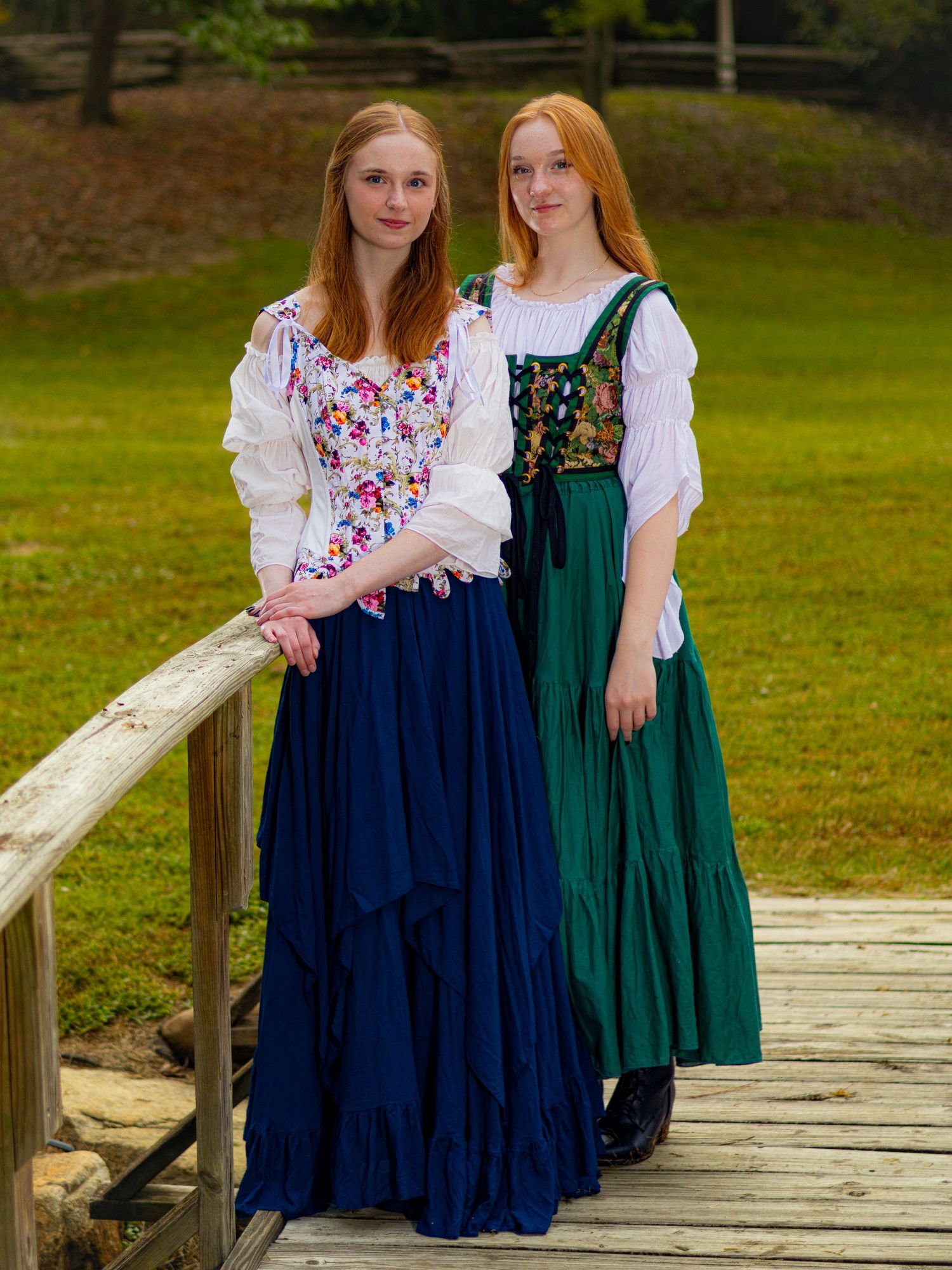 Two young women with red hair wearing colorful folk costumes pose on a wooden bridge outdoors.