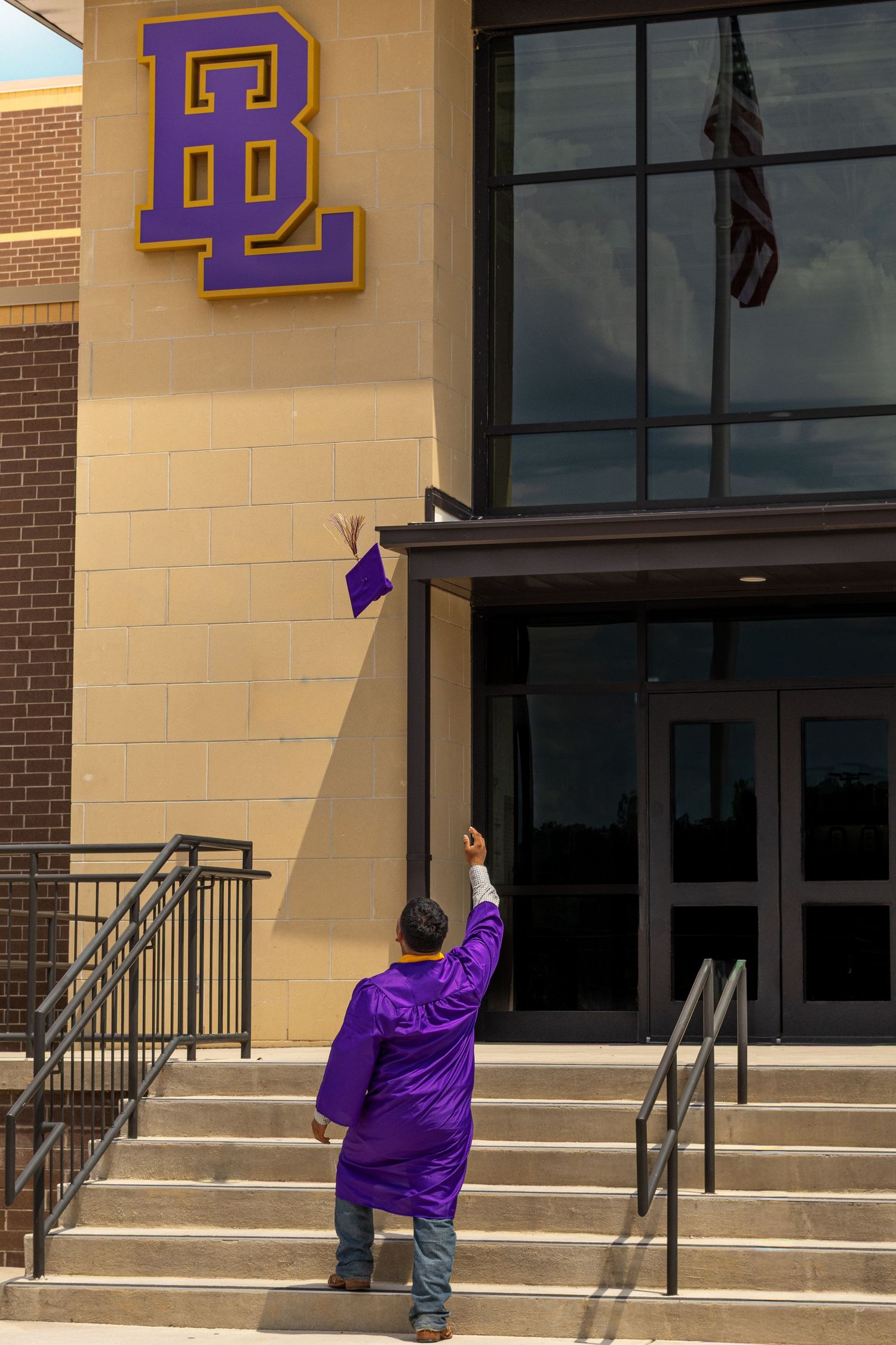 Graduate in purple robe tosses cap outside building with purple and gold BL school logo.