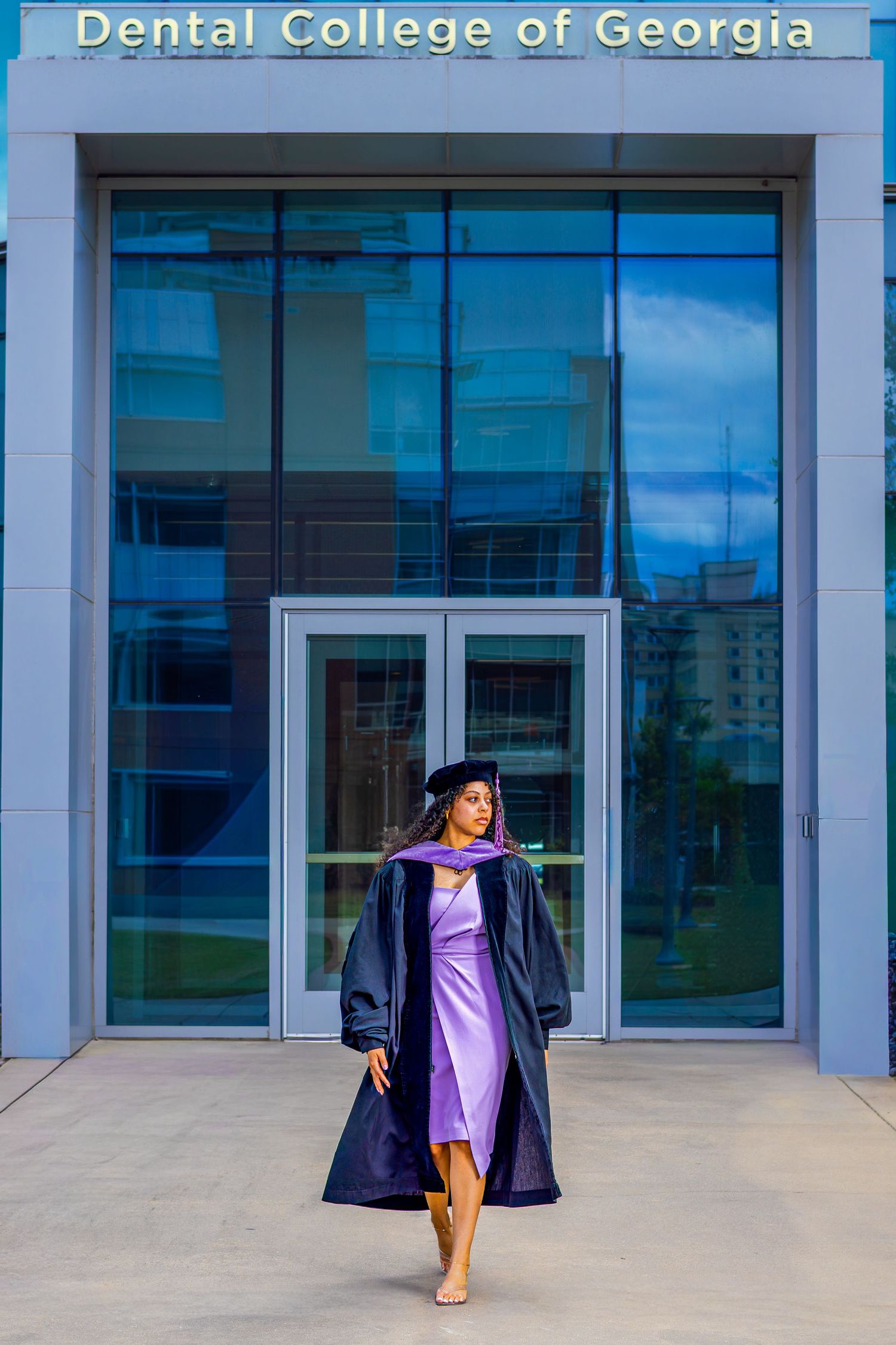 Graduate in cap and gown poses proudly outside the Dental College of Georgia building.