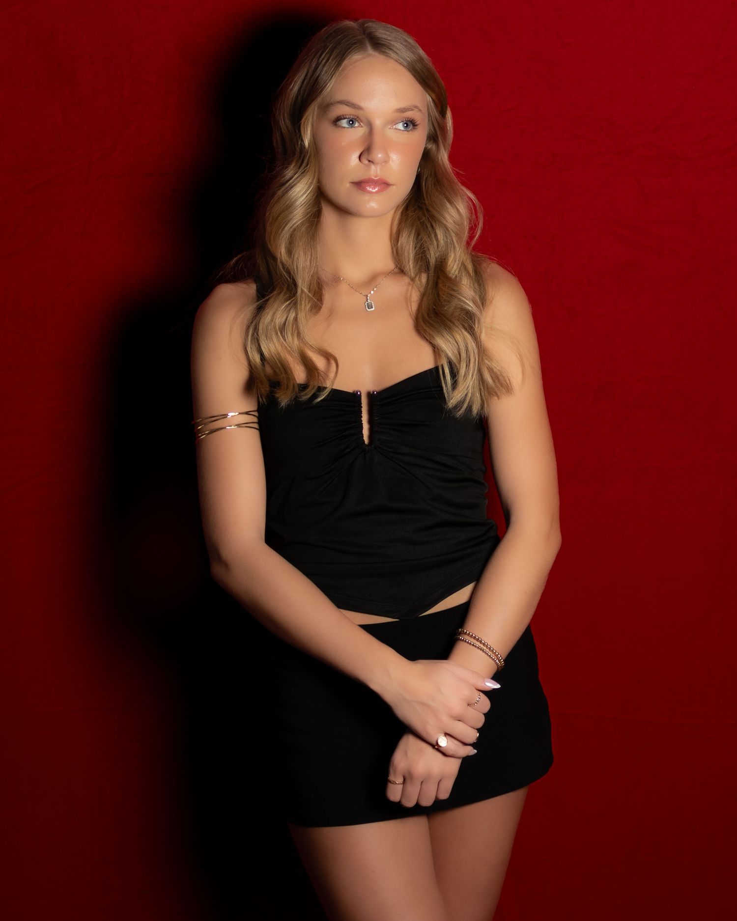Young woman in black crop top and mini skirt poses against a red background wearing gold jewelry.