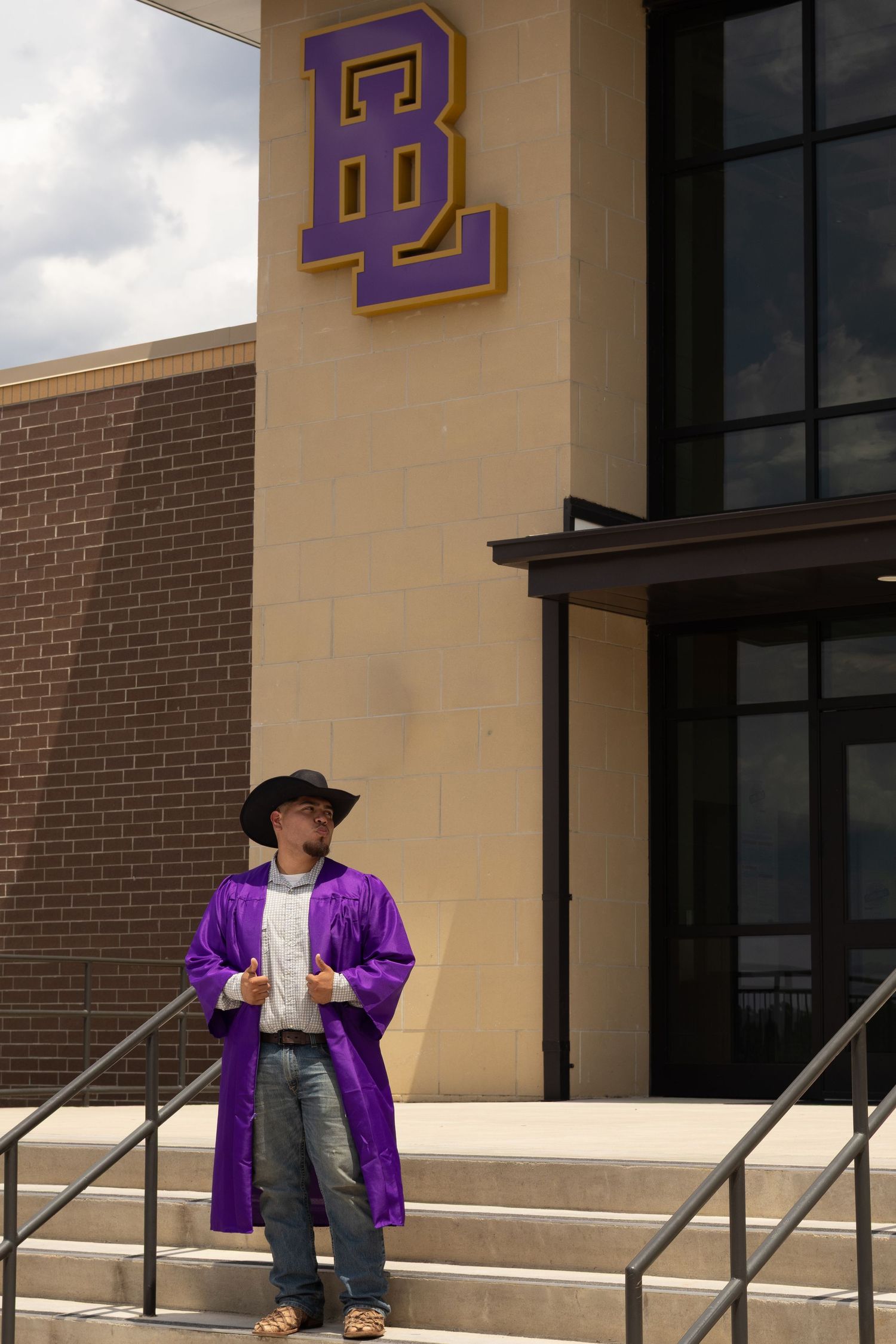 Graduate in purple robe and cowboy hat stands on steps outside a school building with BL logo.
