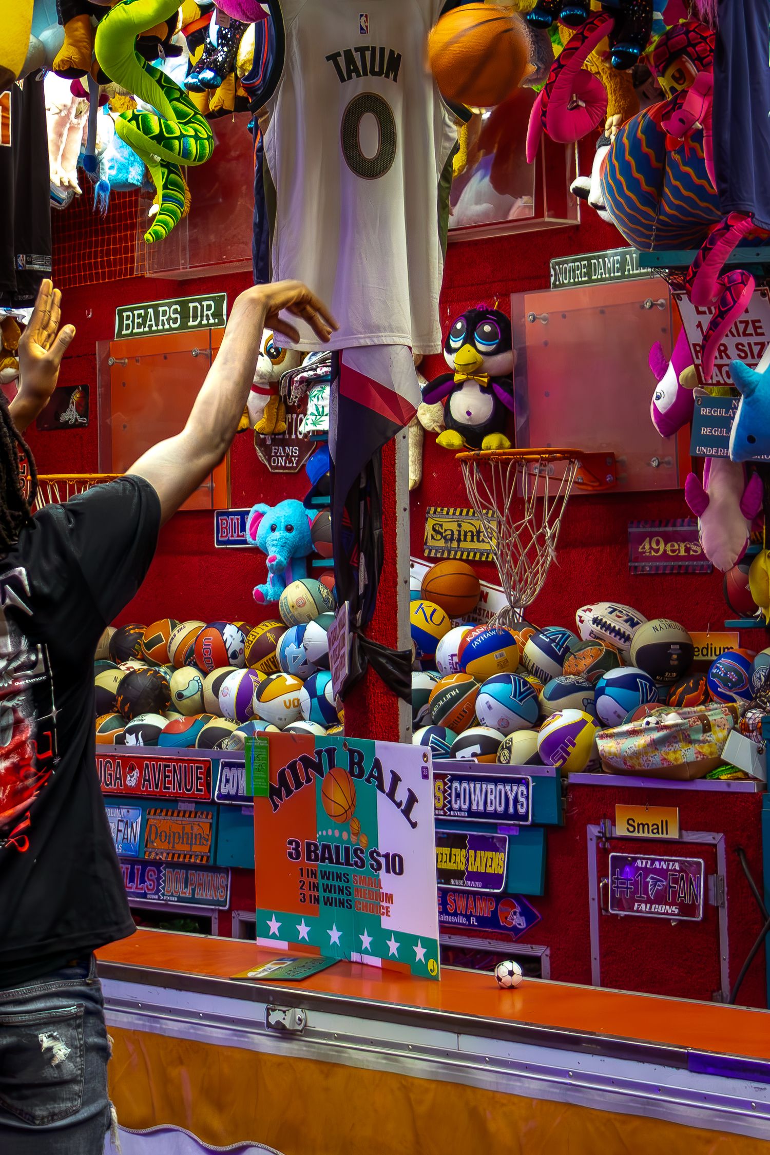 Carnival mini ball toss game booth with colorful stuffed animal prizes and a Jayson Tatum jersey on display.