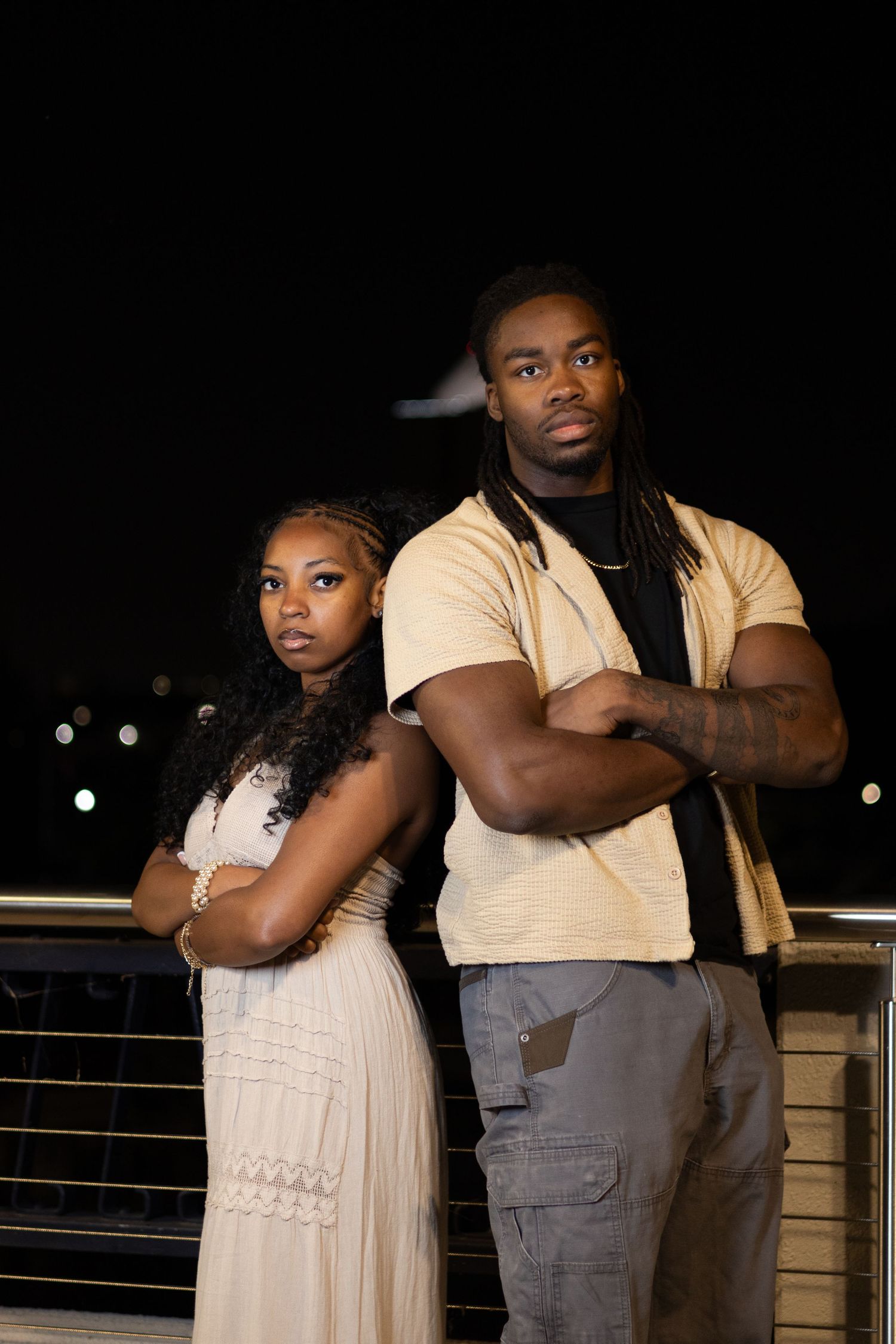 Young Black couple stands back to back with arms crossed in serious pose on nighttime urban bridge.