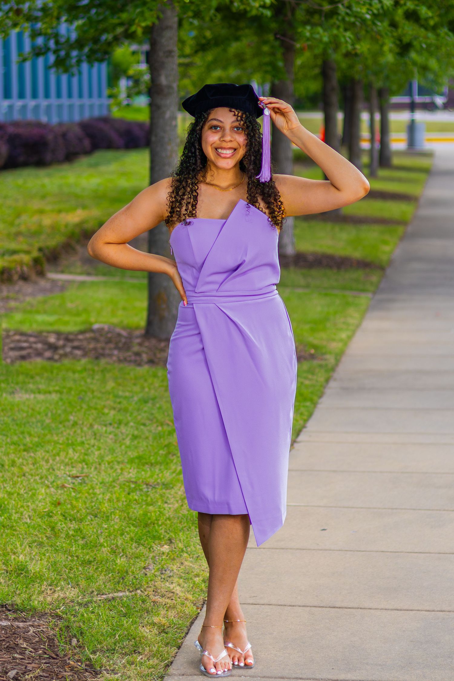 Smiling graduate in purple dress and black cap poses on campus walkway surrounded by green trees.
