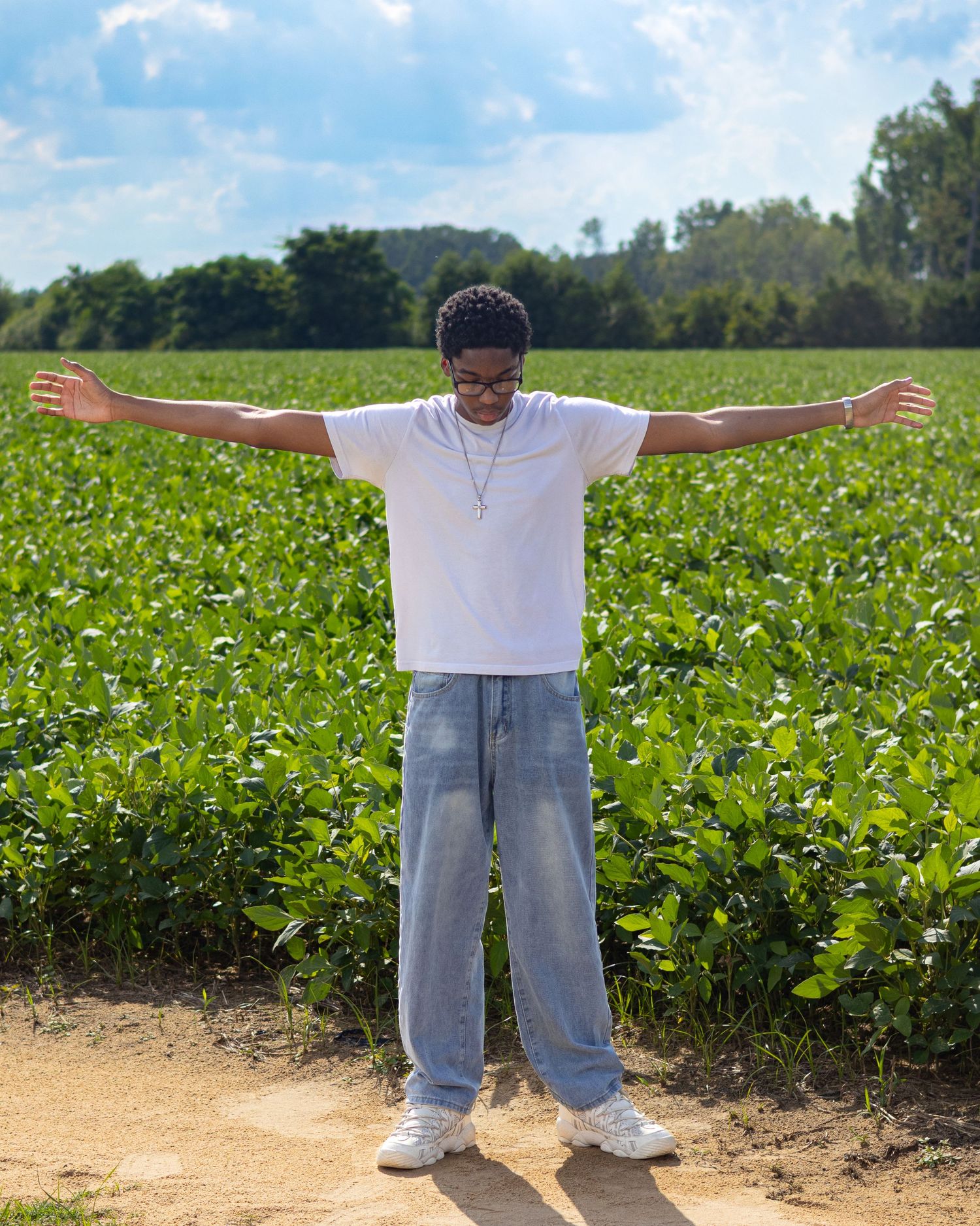 Young man in white tee and jeans stands with arms outstretched in a lush green field under blue sky.