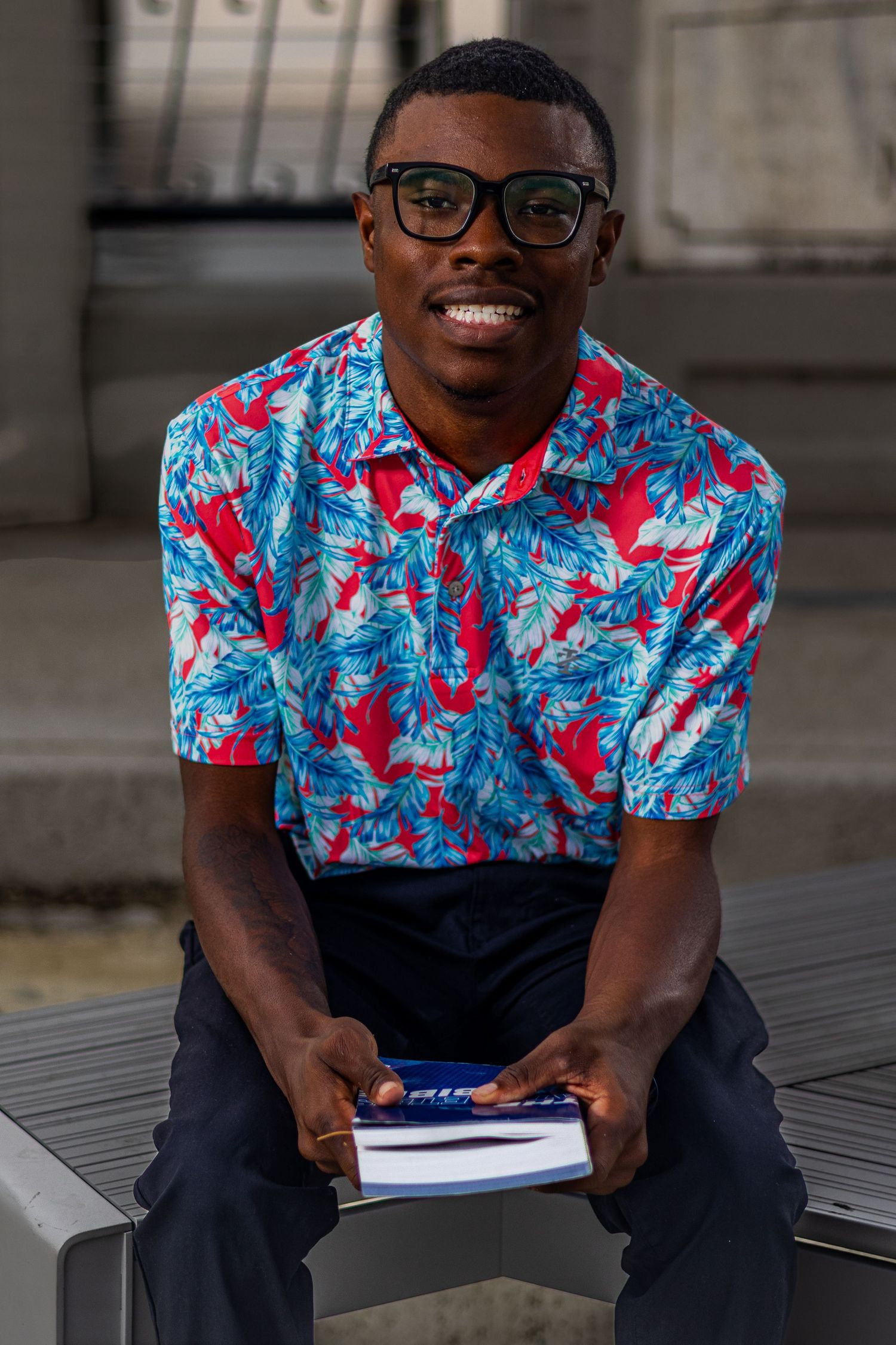 Smiling man in colorful floral shirt and glasses sitting on bench holding a book outdoors.