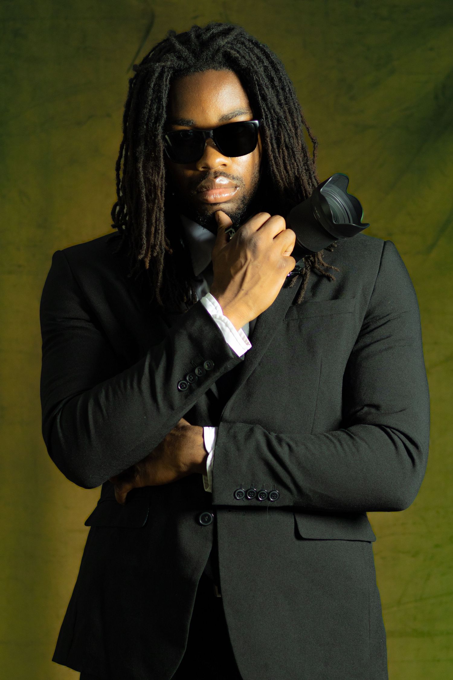 Man with dreadlocks wearing black suit and sunglasses posing against olive green background.