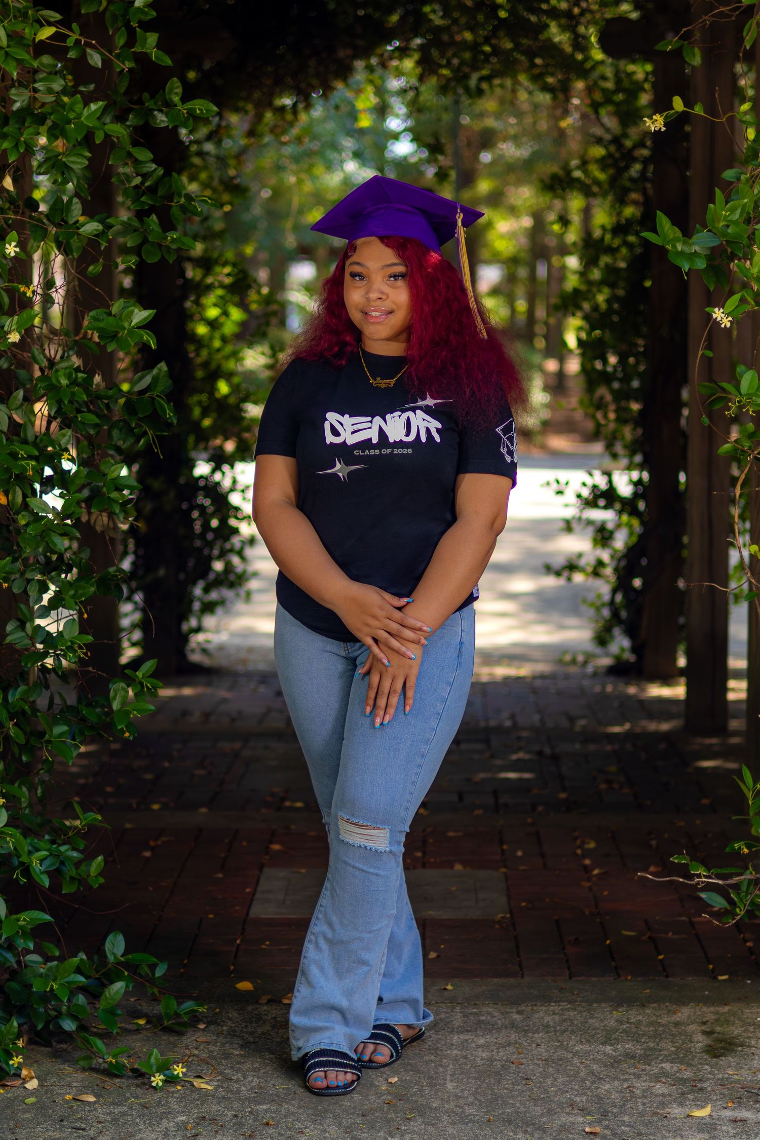 Young woman in purple graduation cap and Senior shirt poses under garden archway for senior photos.