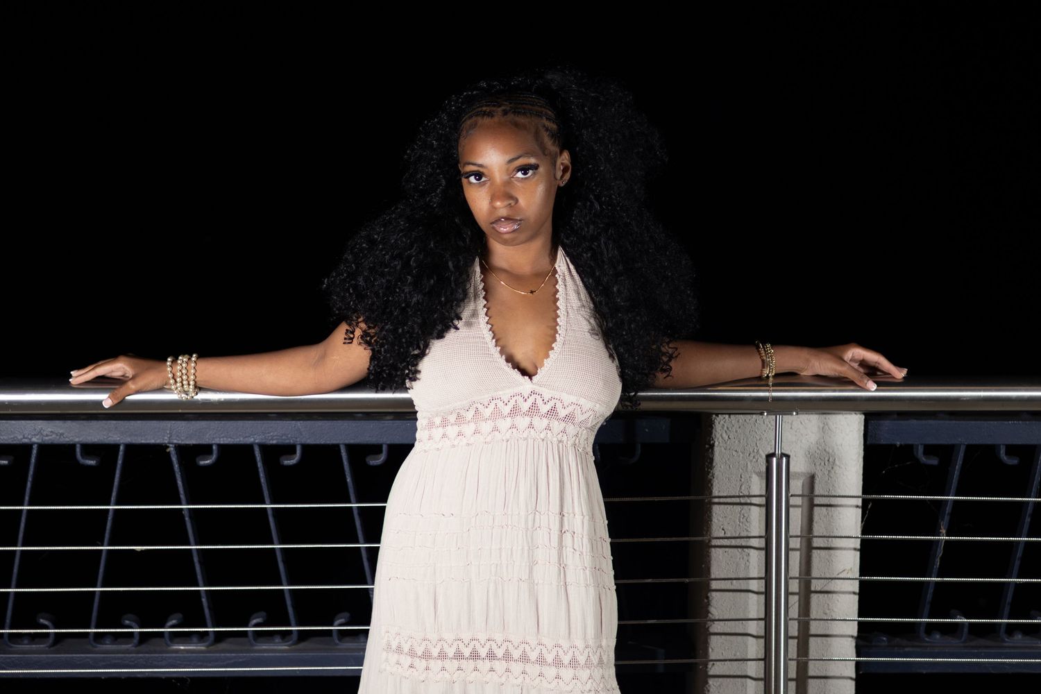 Young Black woman in white dress posing at night on bridge railing.